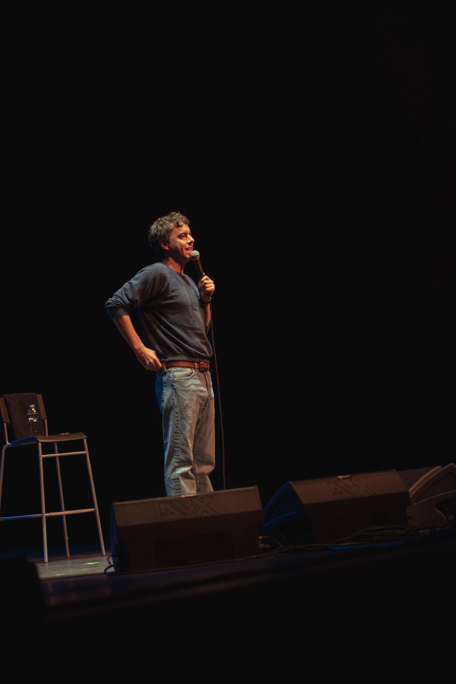 A male performer stands on stage holding a microphone, wearing a dark long-sleeve shirt and jeans, with empty chairs and a water bottle on stage.