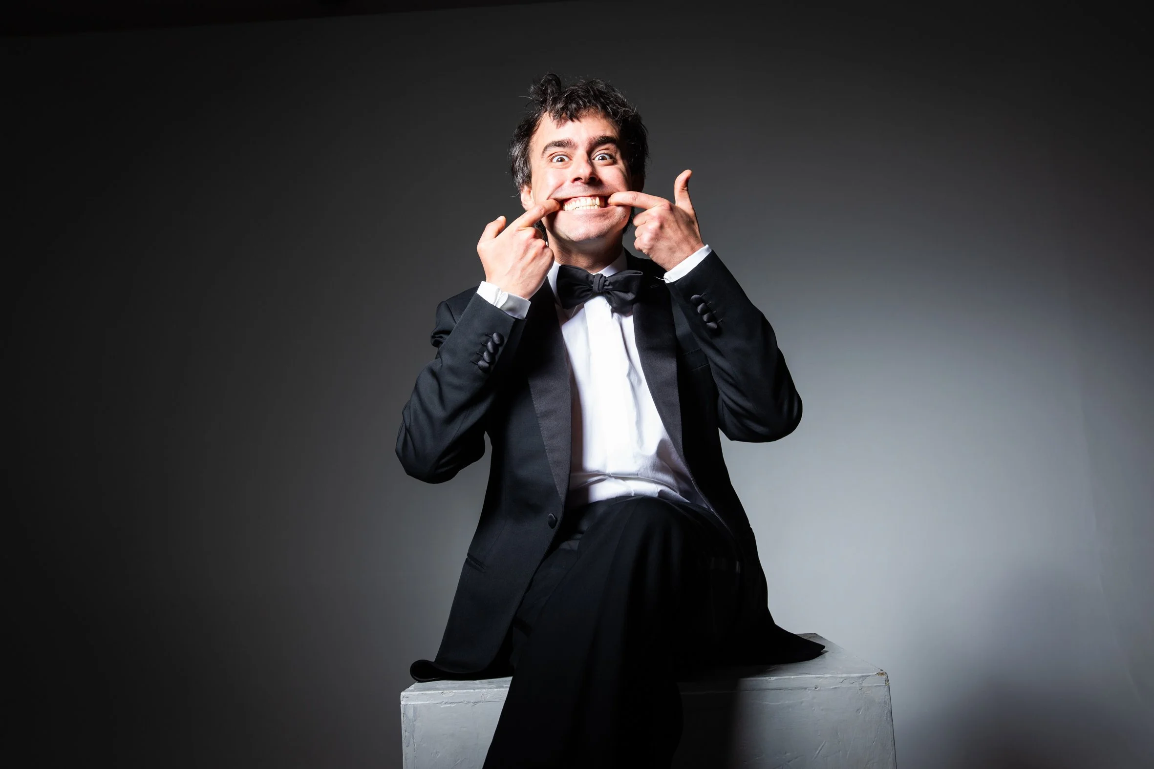 A man in a tuxedo making a silly face by pulling his mouth wide and tugging at his cheeks with his fingers, sitting on a white block against a dark background.