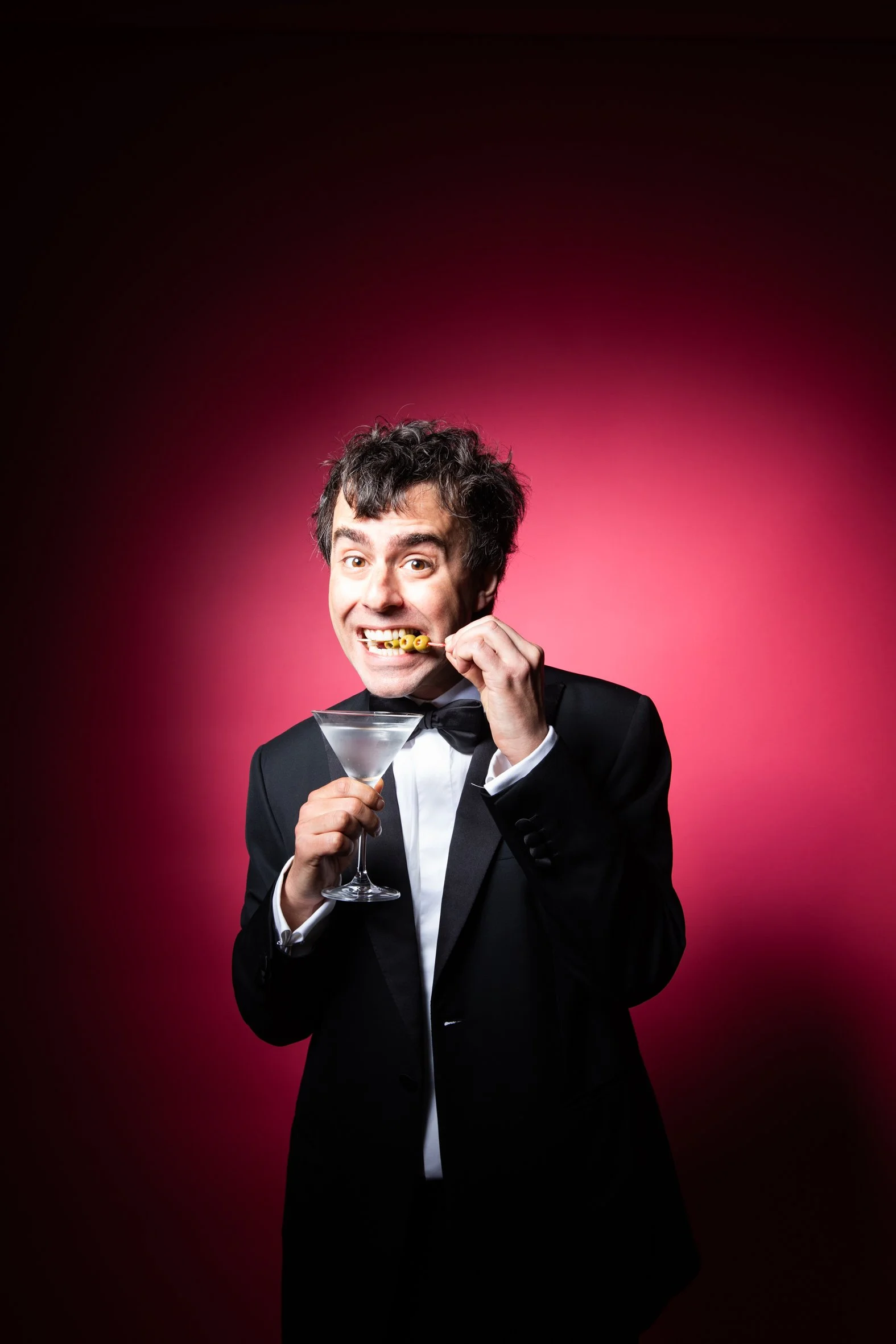 A man in a tuxedo with a bow tie holding a martini glass, biting yellow pearl-like candies on a stick, with a pink and black gradient background.