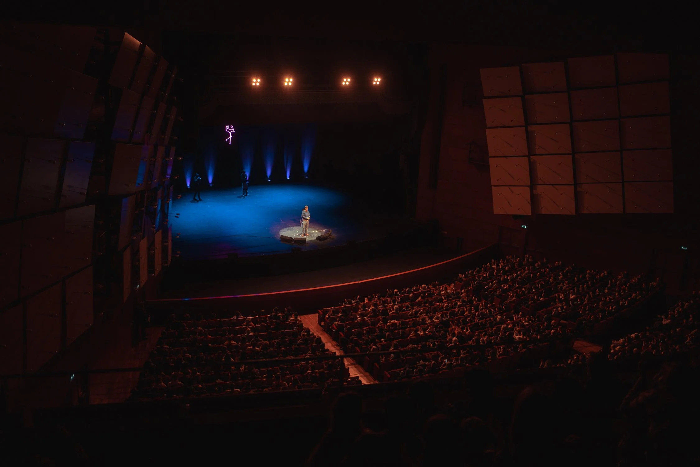 A person speaking on stage in a dark theater with an audience watching, illuminated by blue and pink stage lighting.