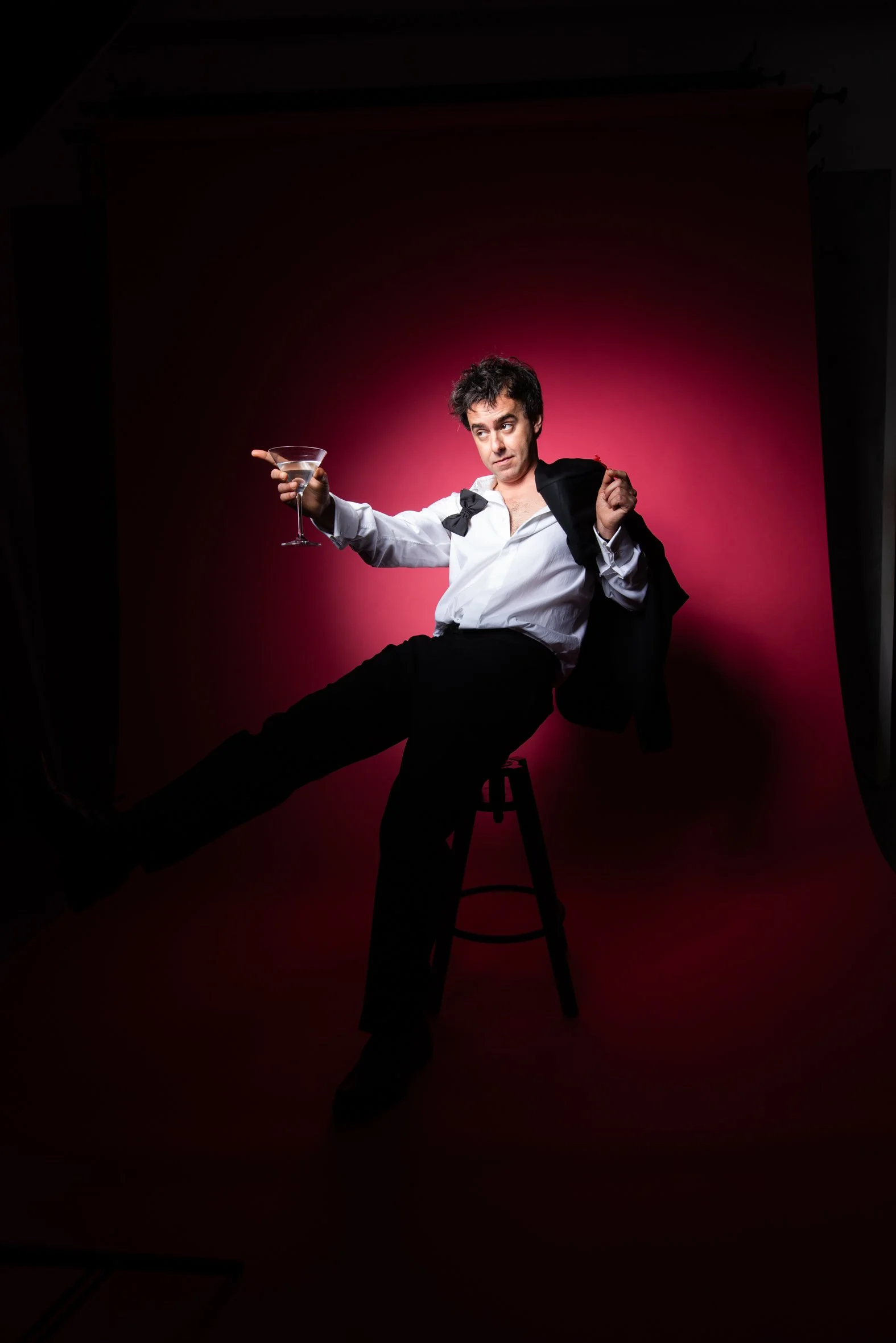 A man with dark, messy hair wearing a white shirt and black pants, sitting on a stool with one leg extended, holding a martini glass in one hand and a black jacket over his shoulder, in a photography studio with a pink backdrop.