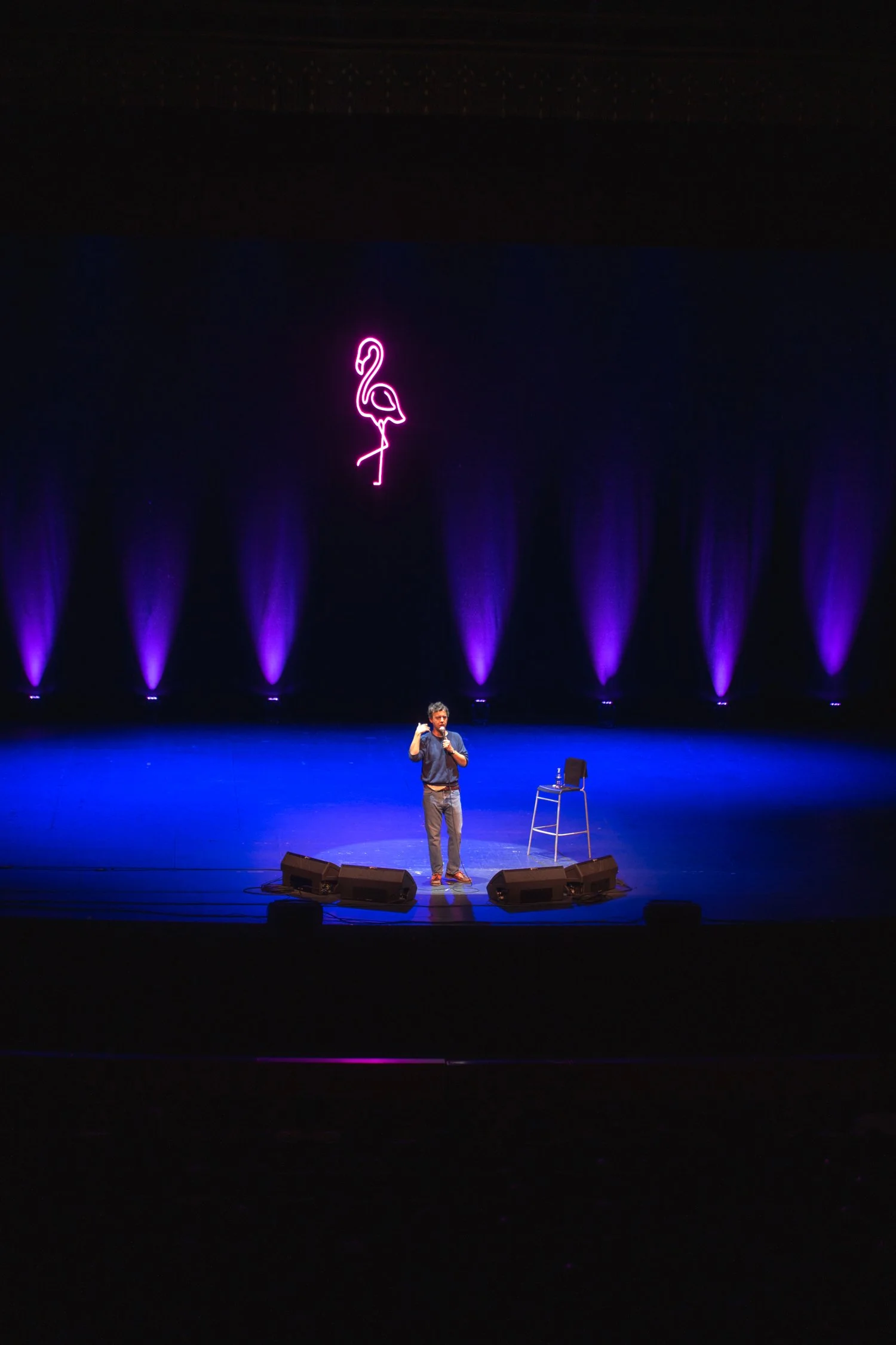 A man is performing on a stage with purple lighting, standing near a stool. A neon pink flamingo sign is hanging above him.