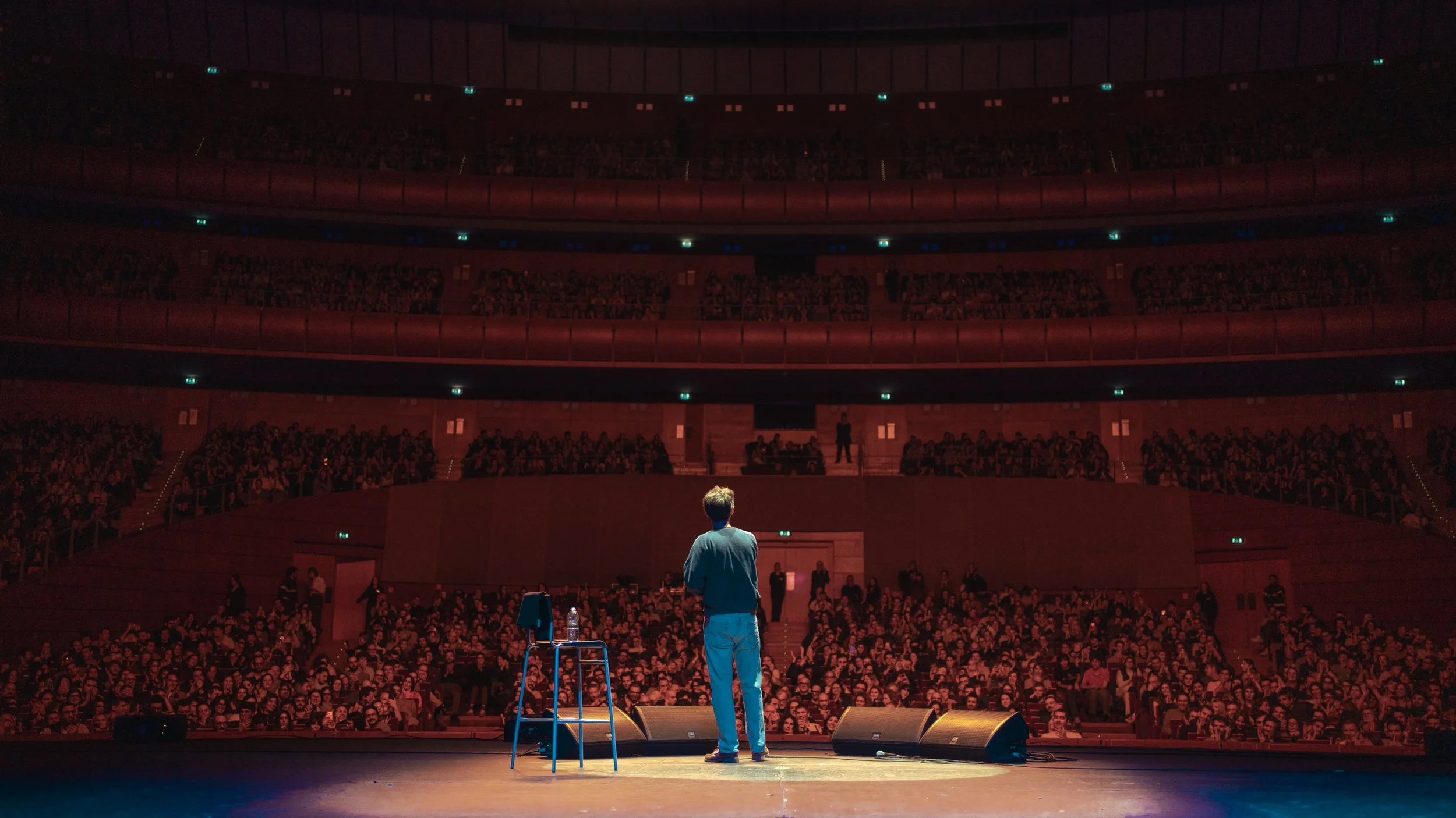 A comedian on stage in front of a large audience in a theater, with a chair, water bottle, and microphone on stage.