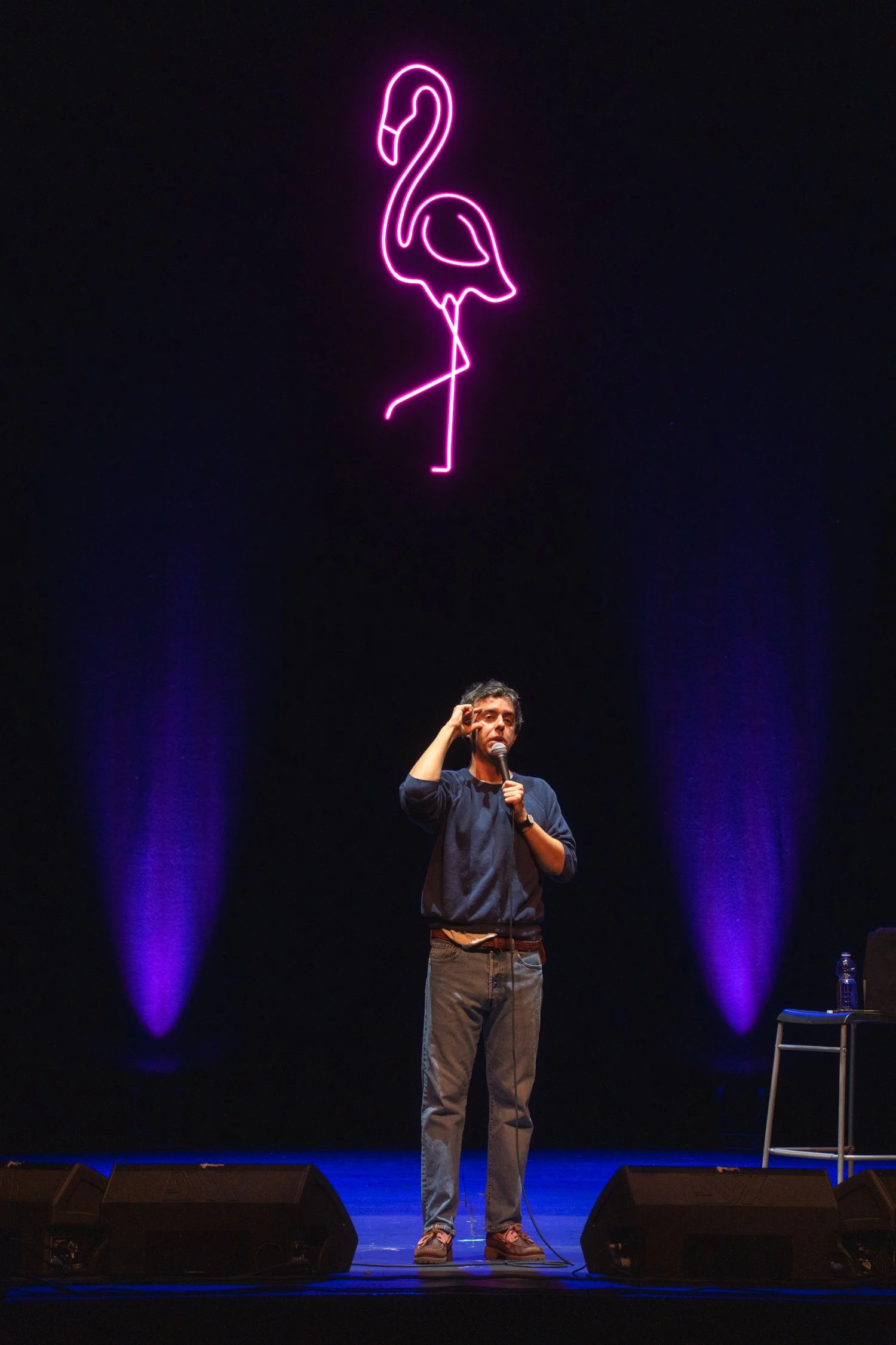 A man holding a microphone on a stage with a neon pink flamingo sign above him.