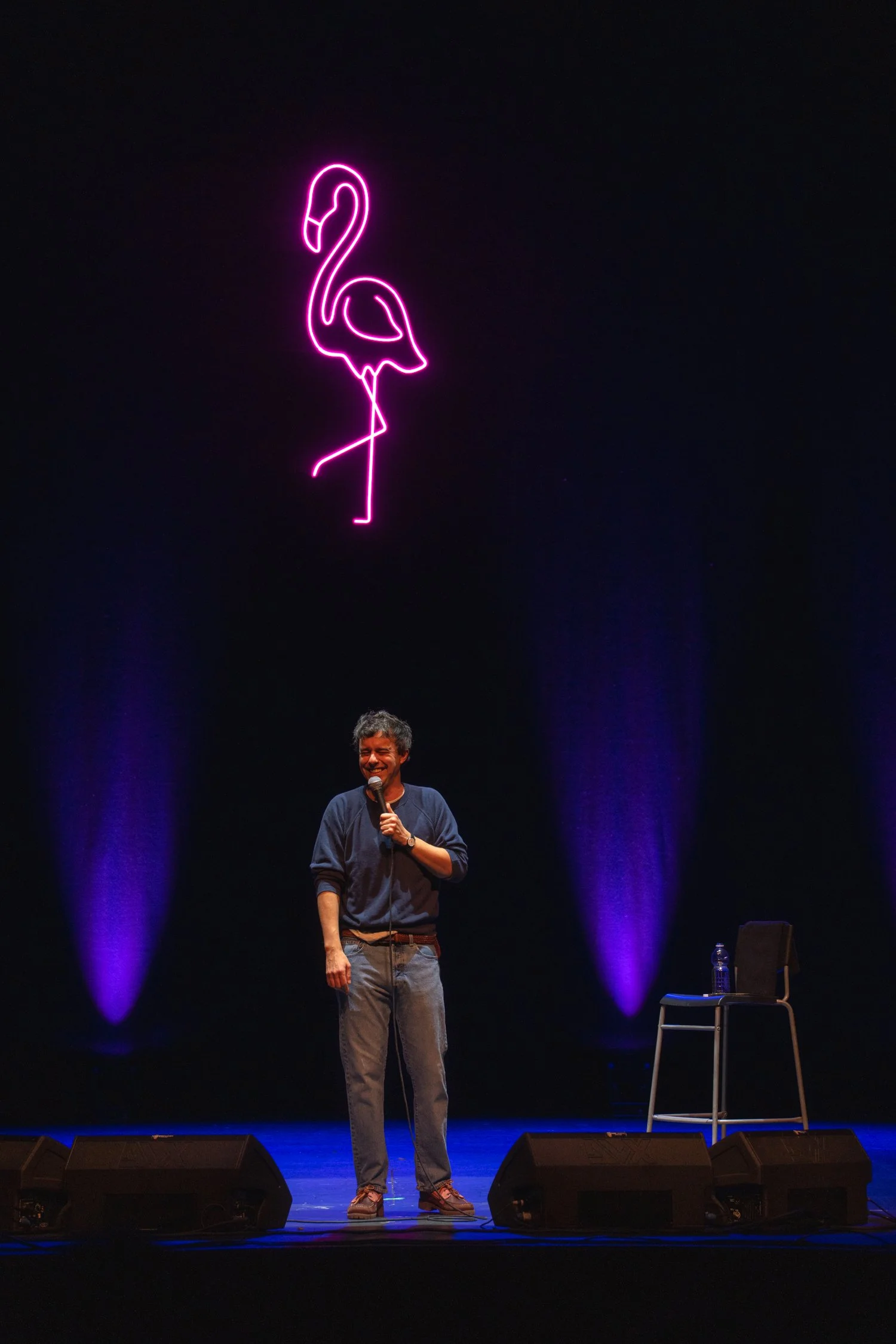 A man standing on a stage holding a microphone, with a neon pink flamingo sign illuminated above him and purple lights highlighting the background.