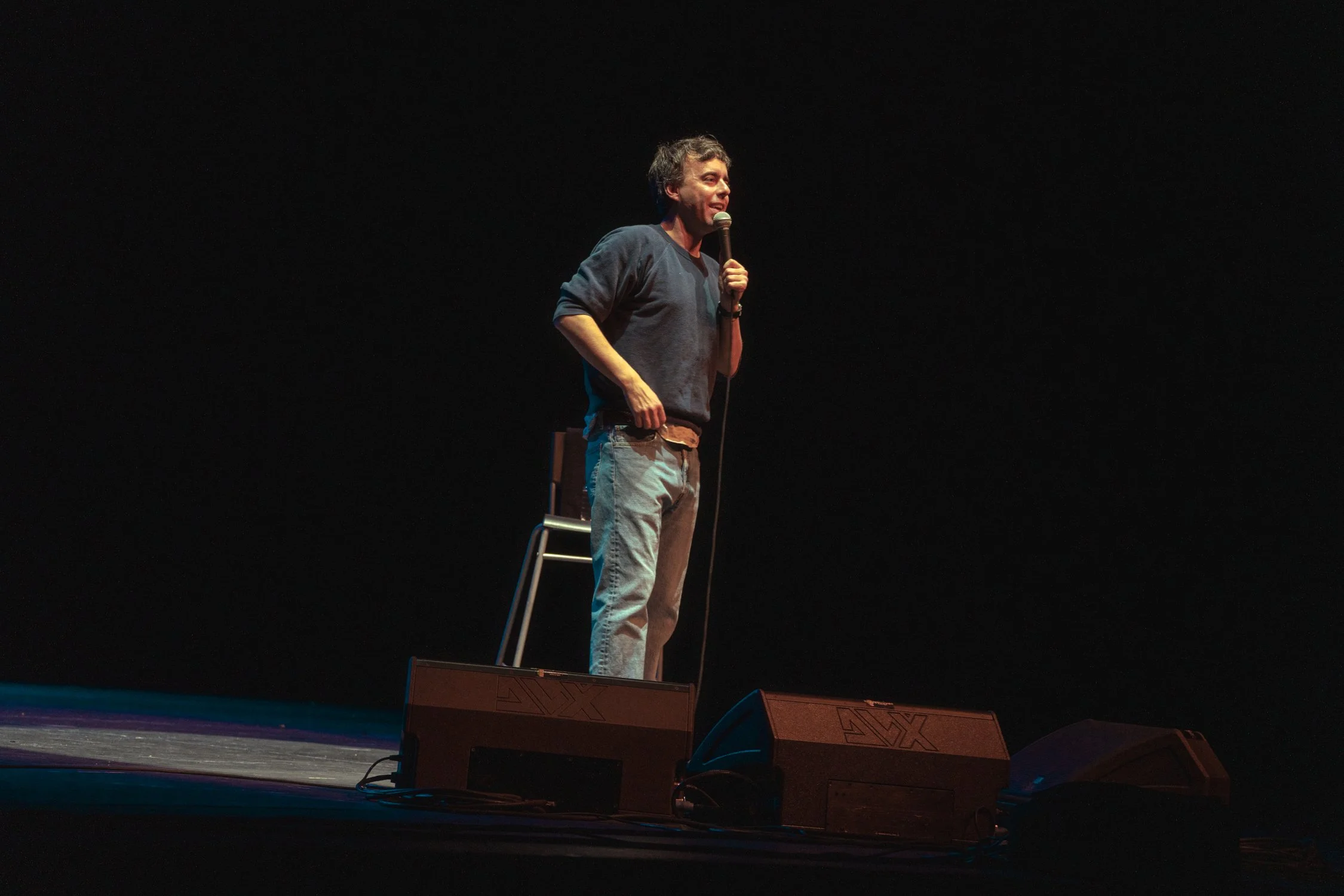 A man performs comedy on stage holding a microphone, standing next to a chair on a dark stage.