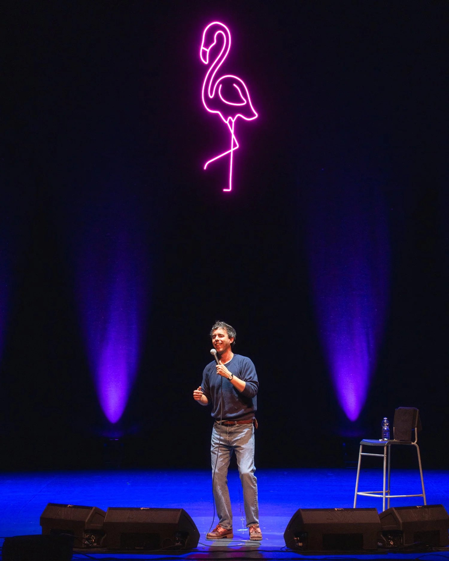 A man standing on stage holding a microphone, smiling, with purple stage lighting and a neon pink flamingo sign hanging above him.