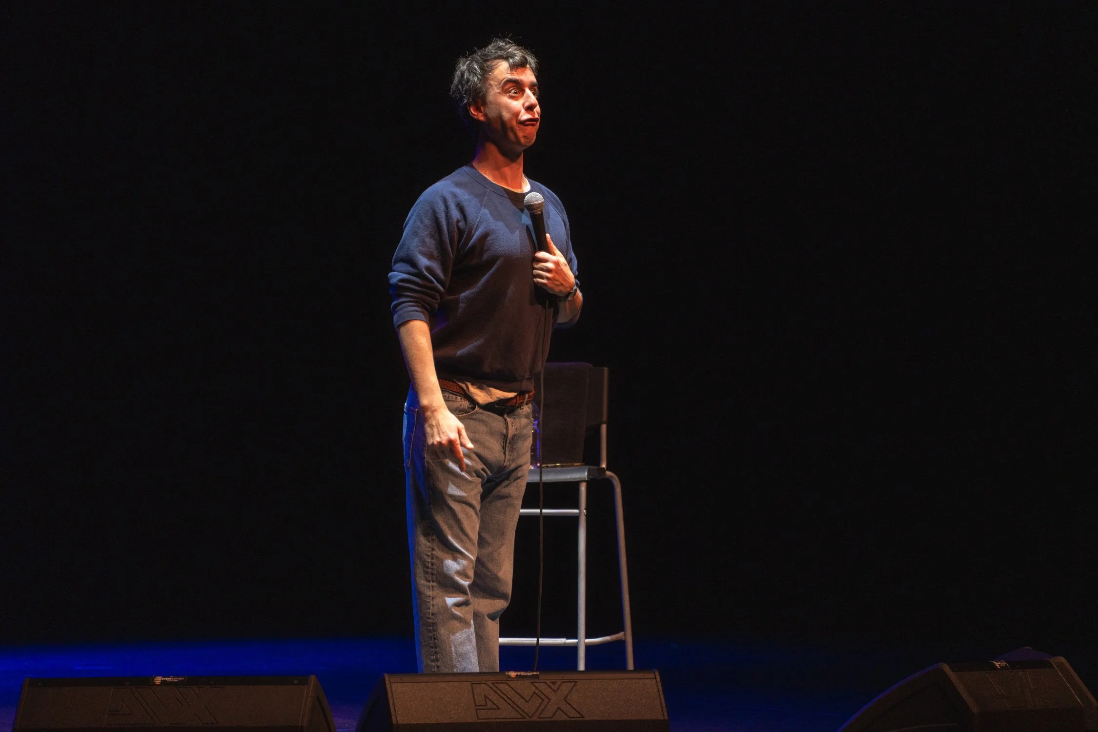 A man standing on a stage holding a microphone, making a funny face, with a chair behind him and speakers at his feet.