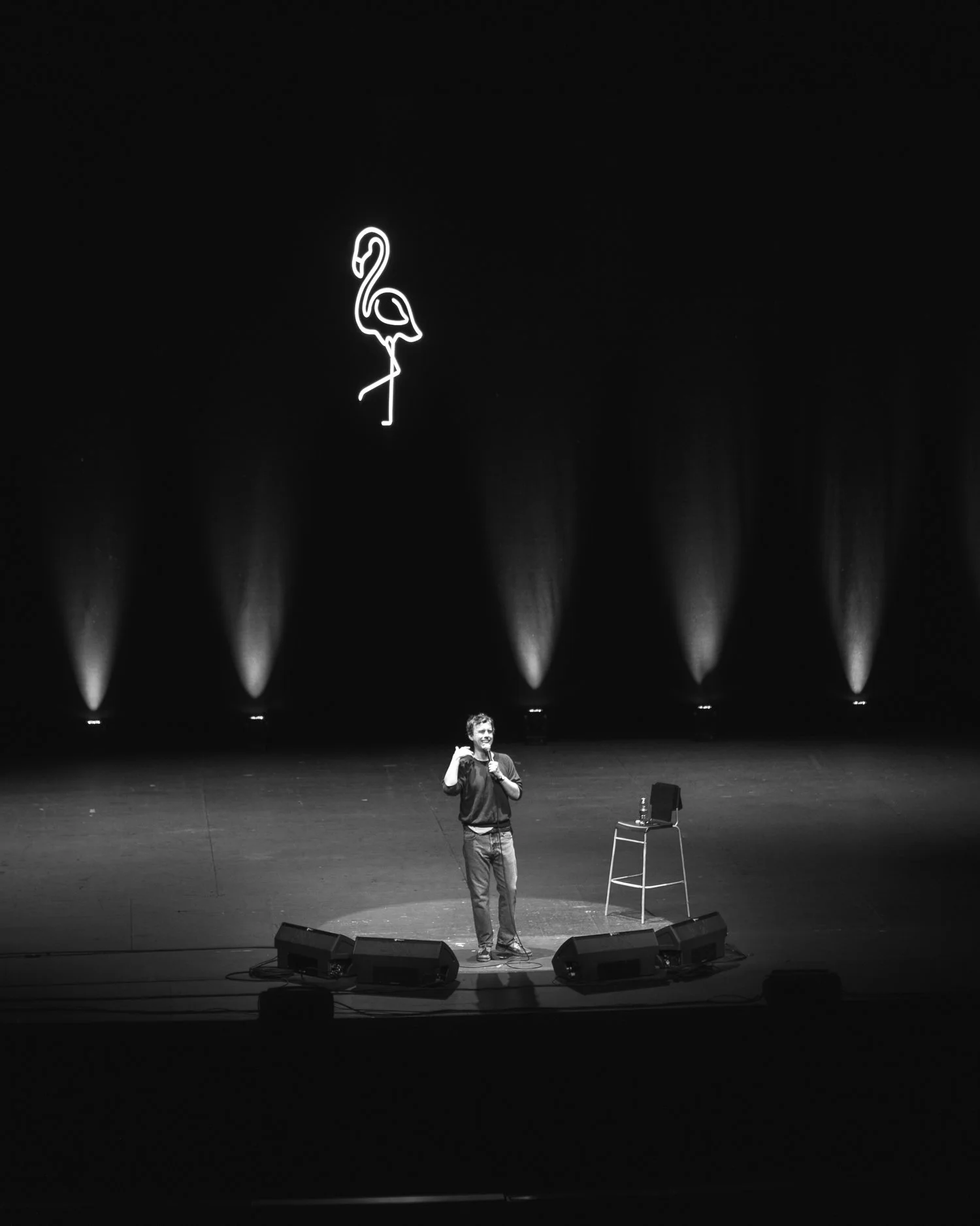 A man stands on a stage with a microphone, smiling and gesturing, with a chair and water bottle beside him. Behind him is a dark backdrop with a neon flamingo sign and pattern lights.