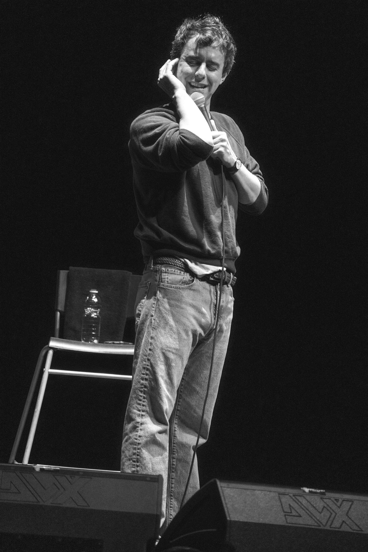 A man standing on a stage, holding a microphone in his right hand, while adjusting an earpiece with his left hand, with a chair, water bottle, and some items on a table behind him.