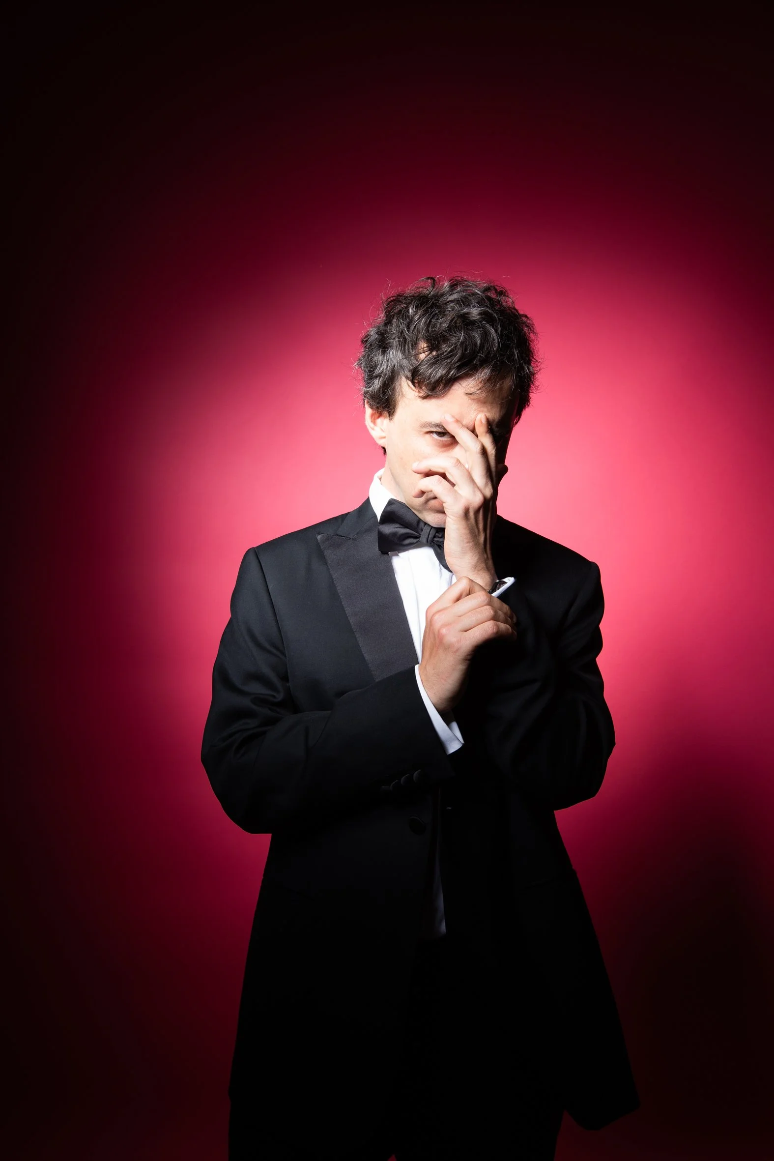 A man in a black tuxedo with a bow tie standing in front of a pink and black gradient background, with his hand partially covering his face in a thoughtful pose.