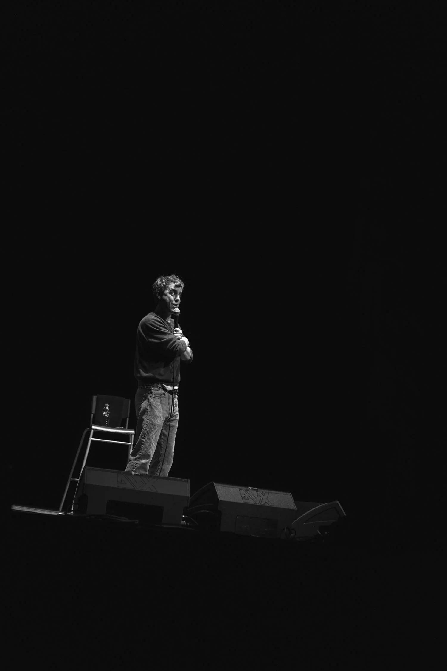 A man standing on a stage, holding a microphone, with a chair and speakers nearby, performing in a dark setting.