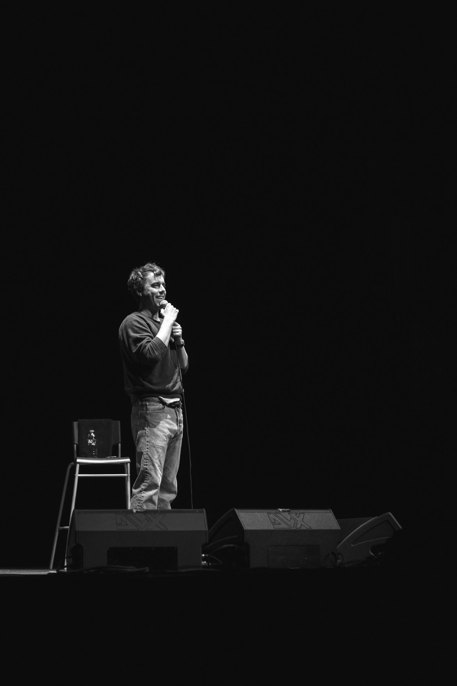 A person on stage holding a microphone, standing next to a chair with a water bottle, in a dark setting.