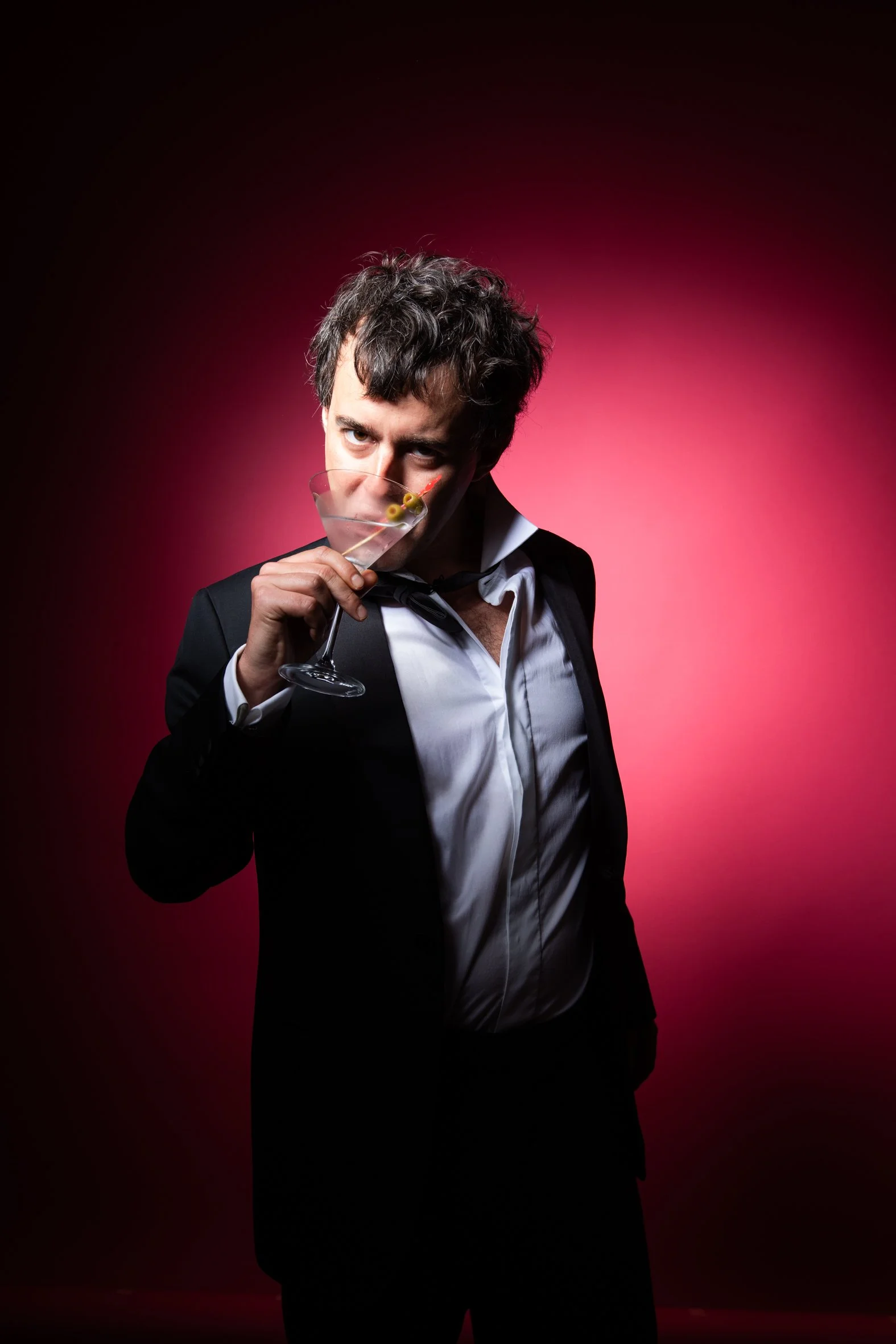 A man in a tuxedo holding a martini glass and looking at the camera with a playful expression.