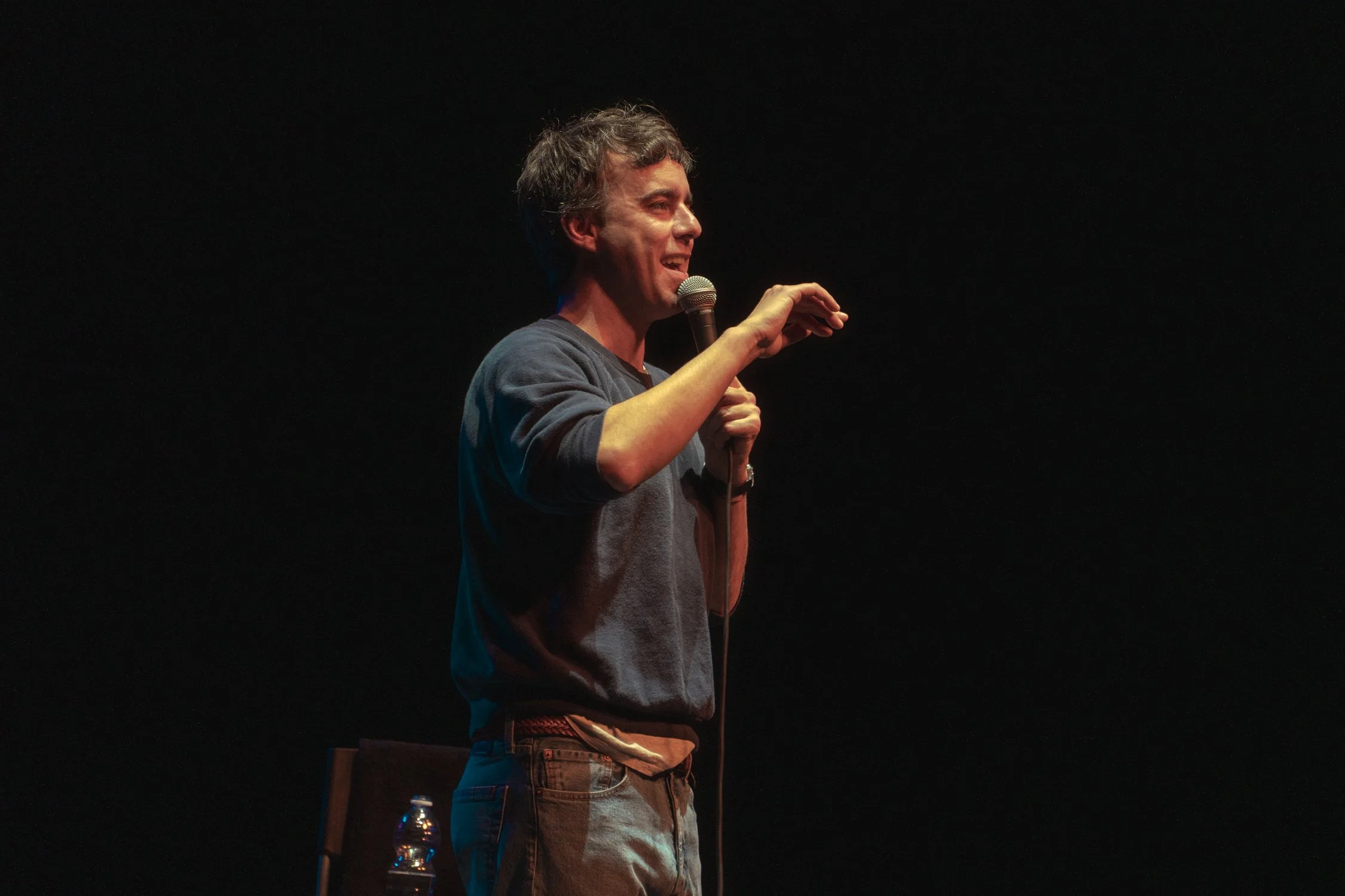A man holding a microphone and laughing, standing against a black background.