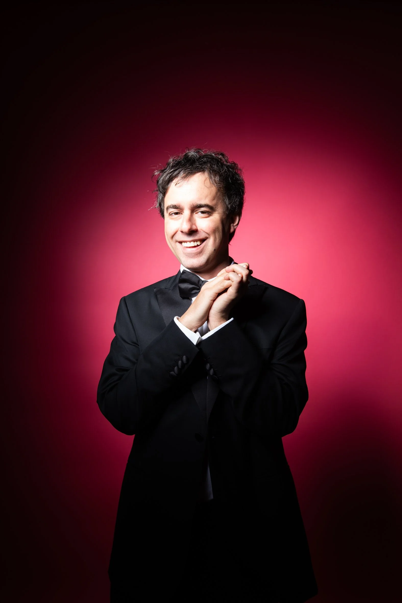 A man in a tuxedo smiling, with hands clasped, against a pink and black gradient background.