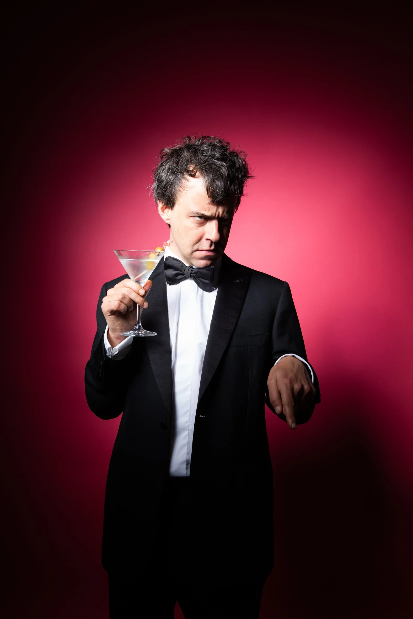 A man in a tuxedo holding a martini glass, looking skeptical against a pink background.
