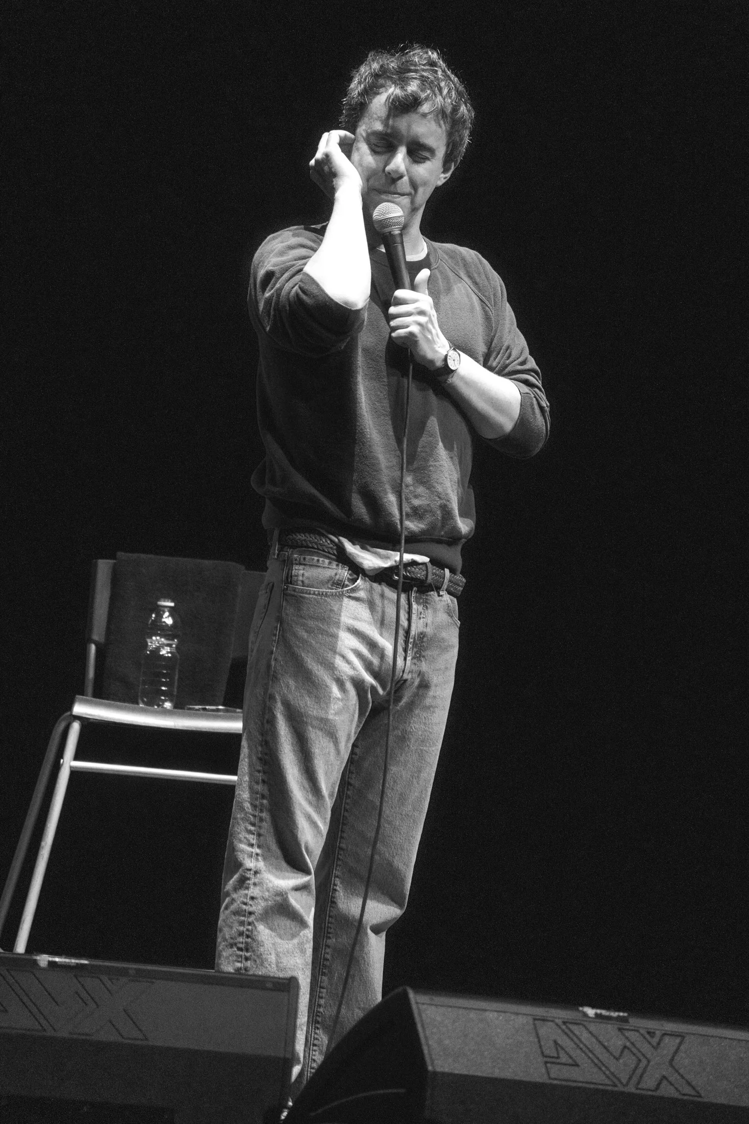 A man with curly hair is standing on stage, holding a microphone close to his mouth. He is wearing a long-sleeved shirt and jeans, with his eyes closed or looking down. There is a chair with a water bottle behind him and speakers in front of him.