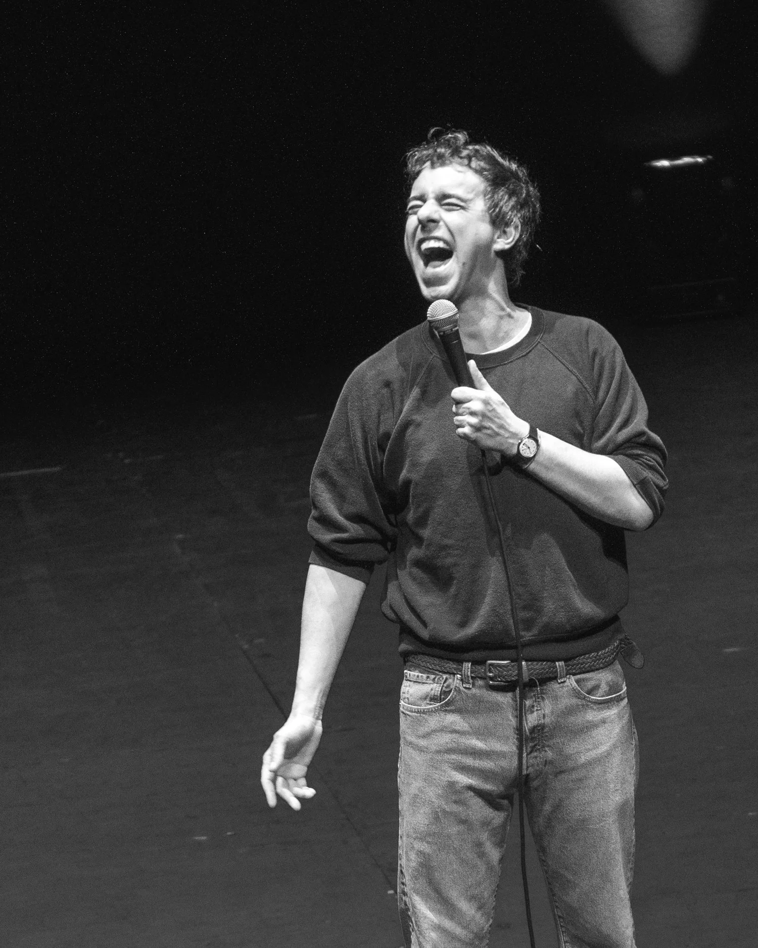 Black and white photo of a young man with curly hair, laughing or singing, holding a microphone in his right hand and wearing a watch and casual clothing, standing on stage.