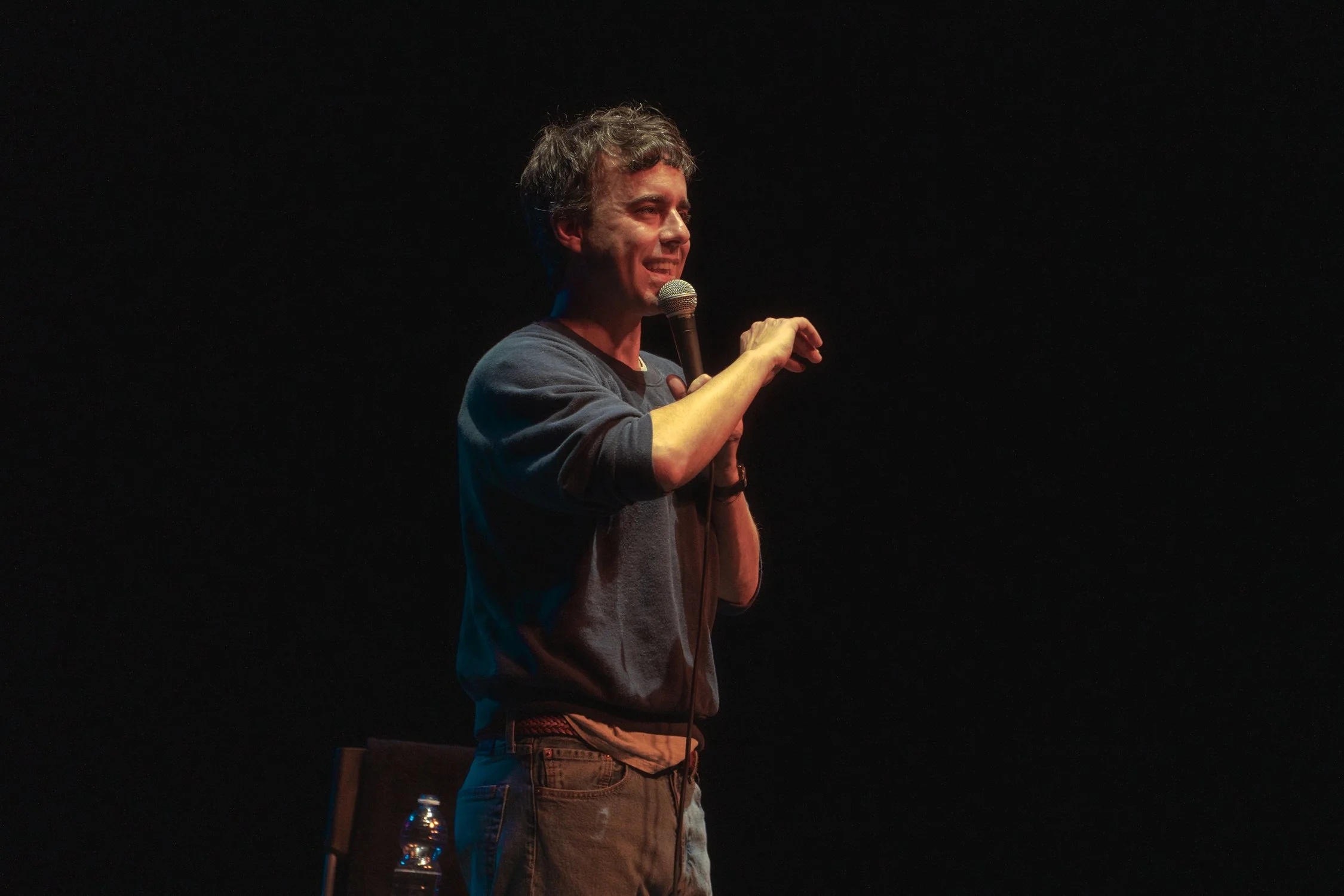 A man with curly hair holding a microphone, smiling, and standing on a dark stage with a black background.