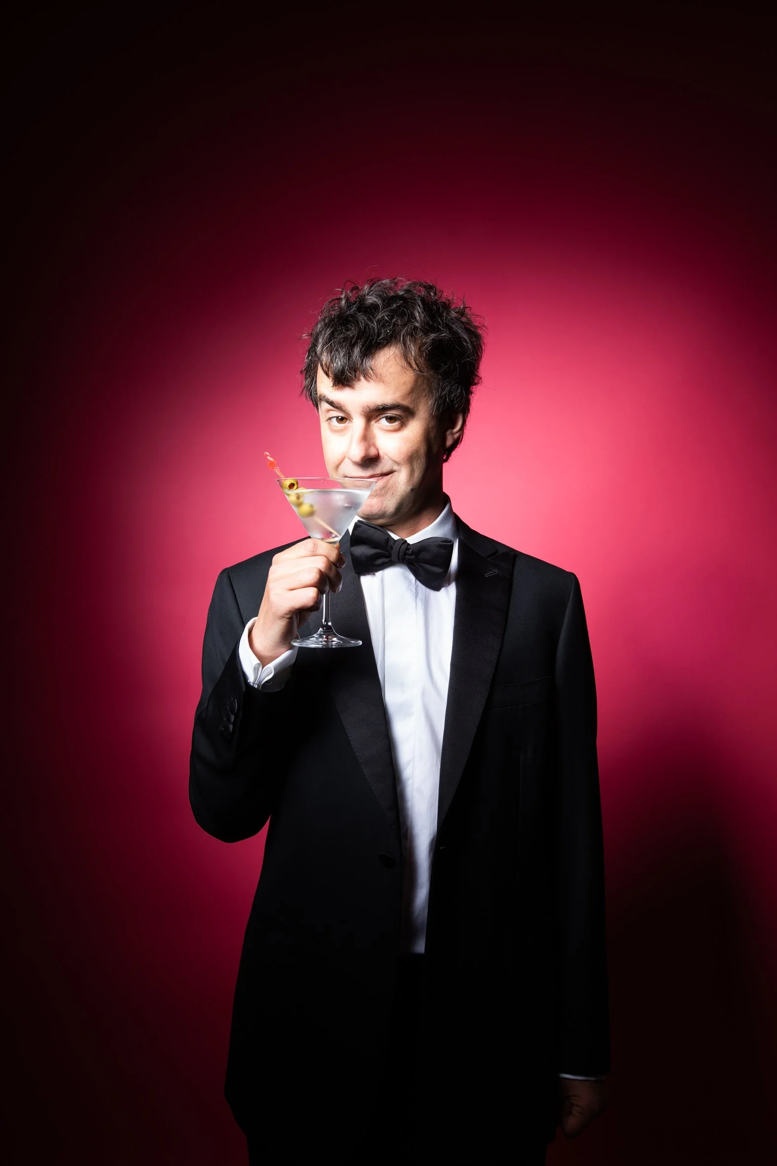 Man in tuxedo holding a cocktail glass against a pink gradient background.