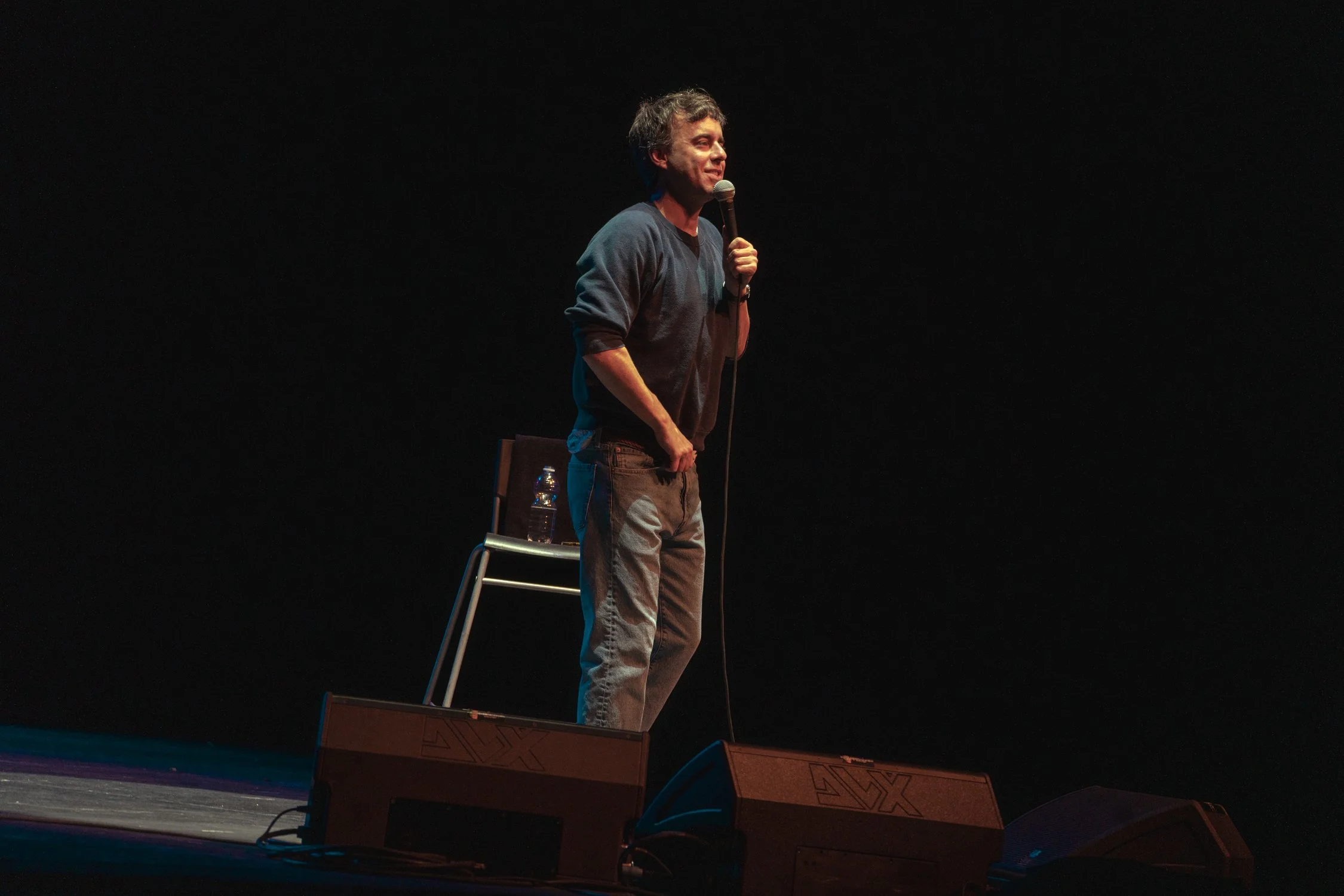 A man stands on a stage holding a microphone, wearing a gray sweater and jeans, with a chair and water bottle behind him, against a dark background.