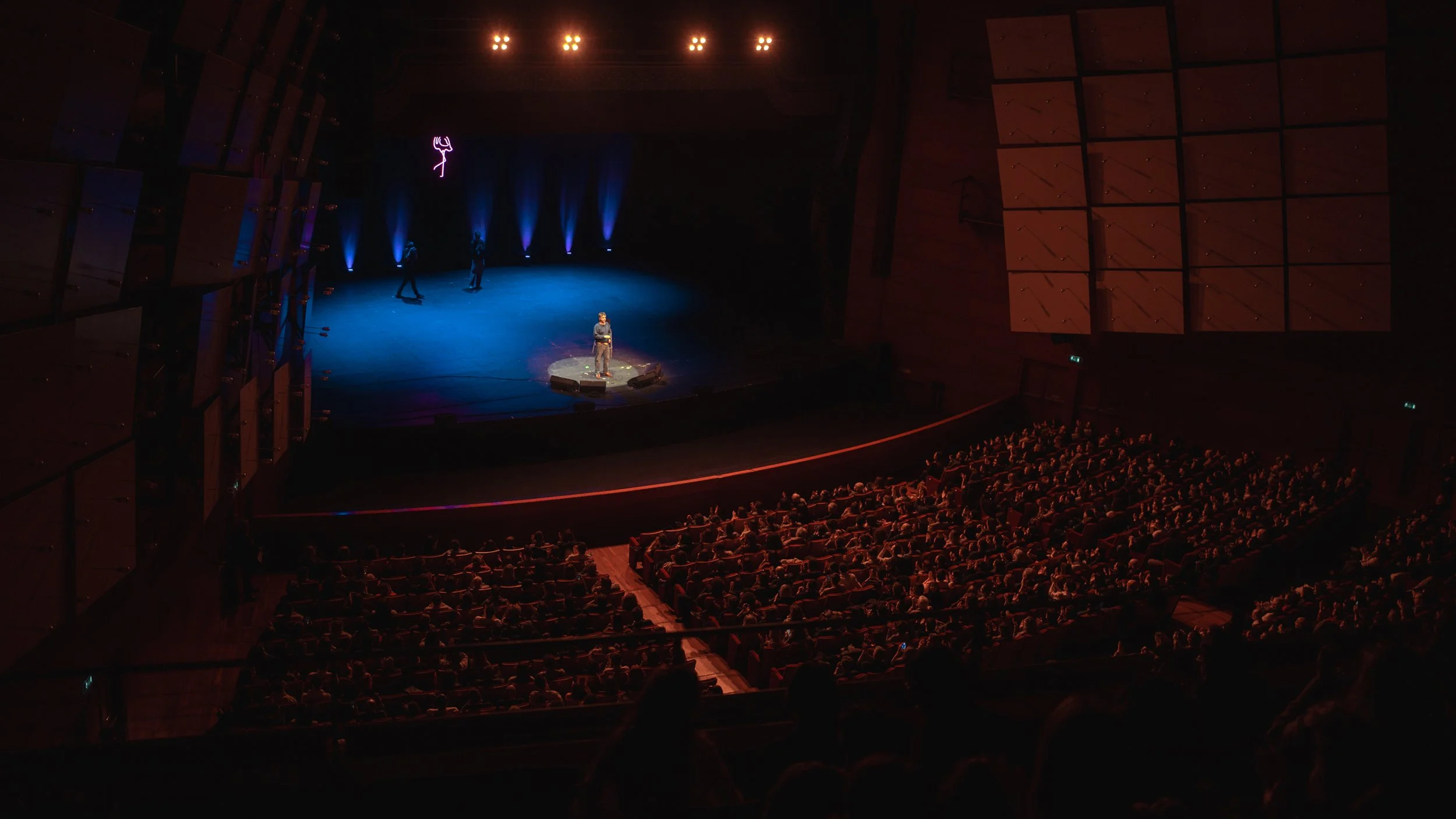 A person is performing on a stage in front of an audience at a theater or concert hall. The stage is lit with blue and purple lighting, and there's a neon pink silhouette of a figure hanging above. The audience is seated in rows, watching the perform