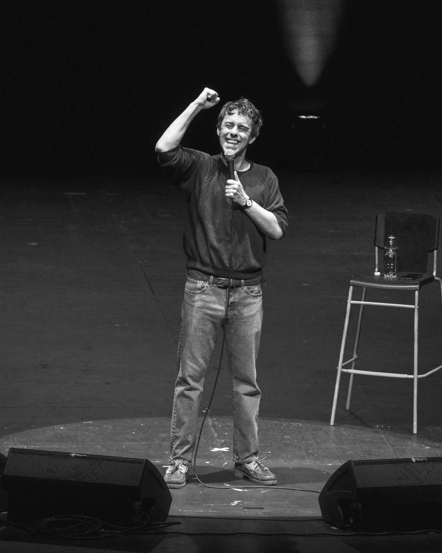 A man standing on stage, holding a microphone, smiling, and raising his fist in the air. There is a chair with a water bottle on it to his right and two speakers at his feet.