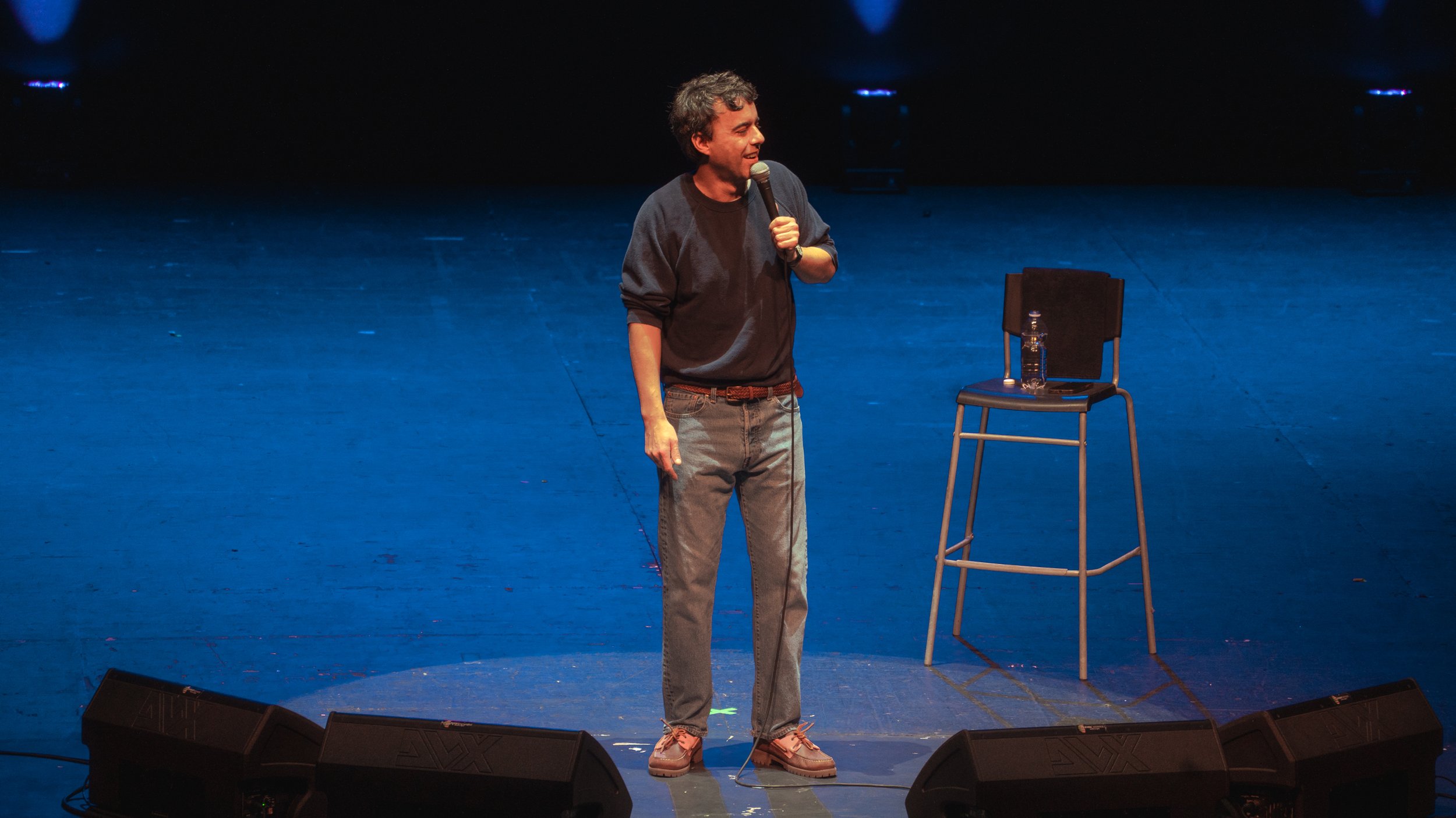 A man performs stand-up comedy on a stage, holding a microphone, with a chair and water bottles behind him, under stage lights.
