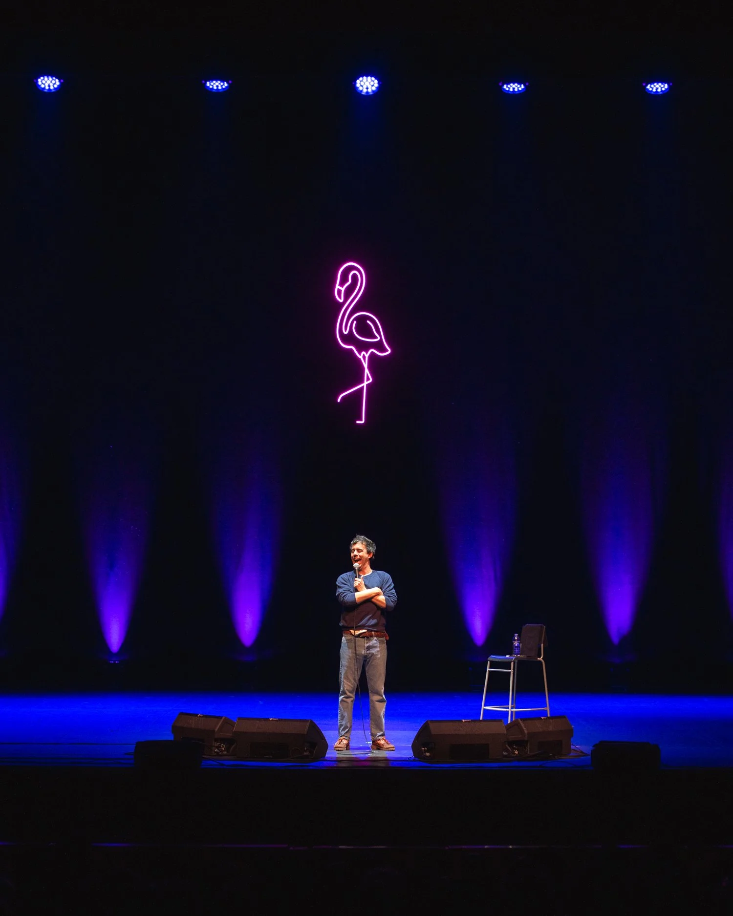 A man standing on a stage holding a microphone, with a neon pink flamingo sign hanging above him. The stage is illuminated with purple lights, and there is a chair with a water bottle on it to the side.