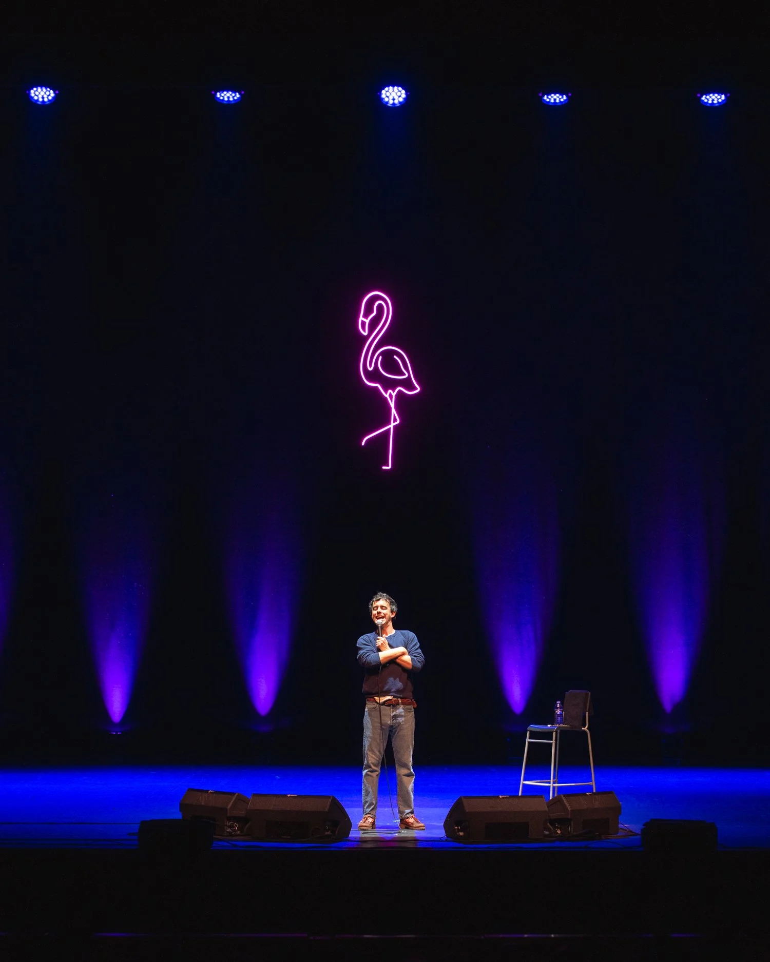 Stand-up comedian on stage with a microphone, purple flamingo neon sign hanging above, purple stage lights, and a chair with a water bottle on it to the right.