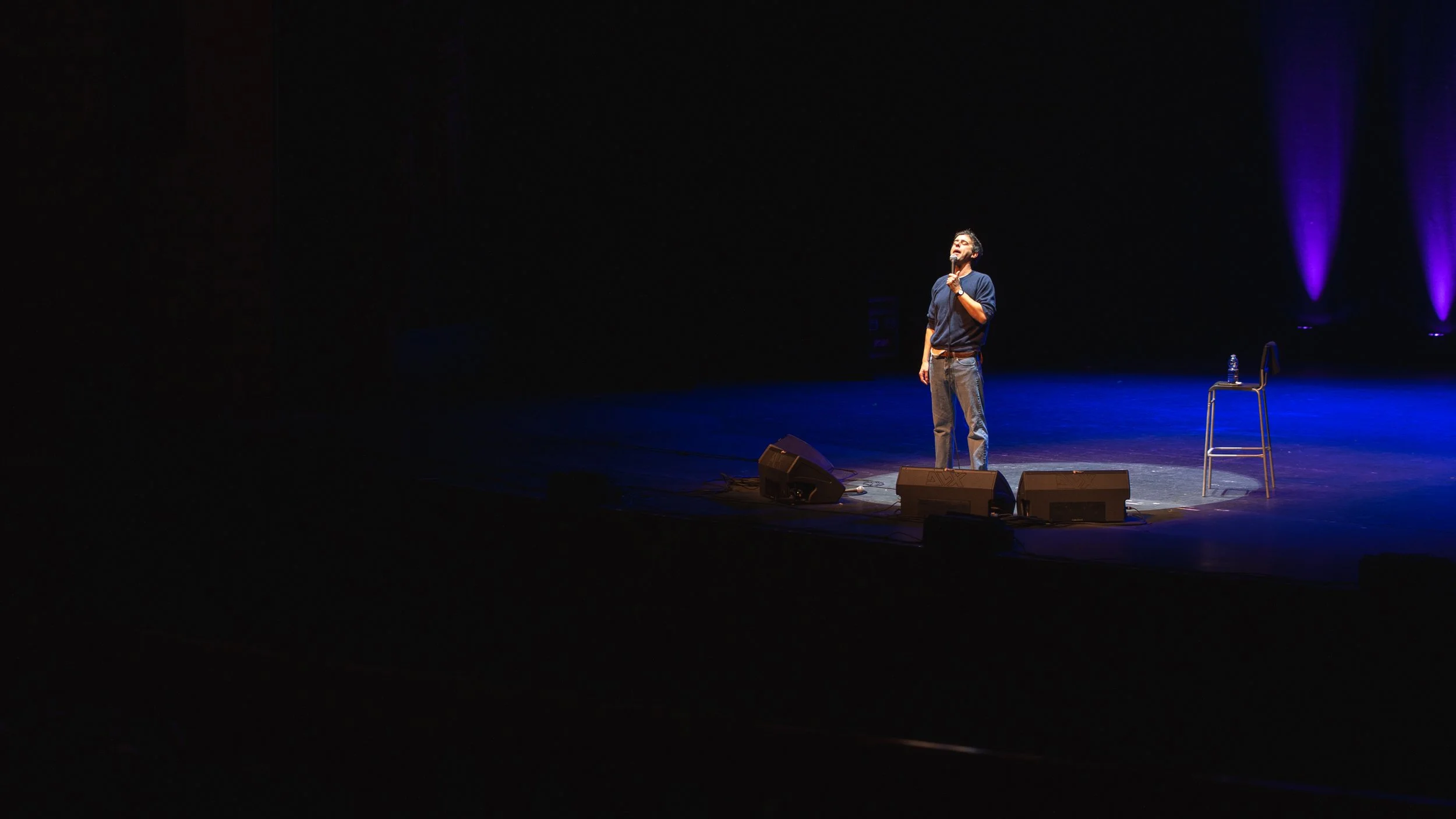 A person stands on stage holding a microphone, with a chair and a water bottle beside him. The stage is dimly lit with purple lights in the background.