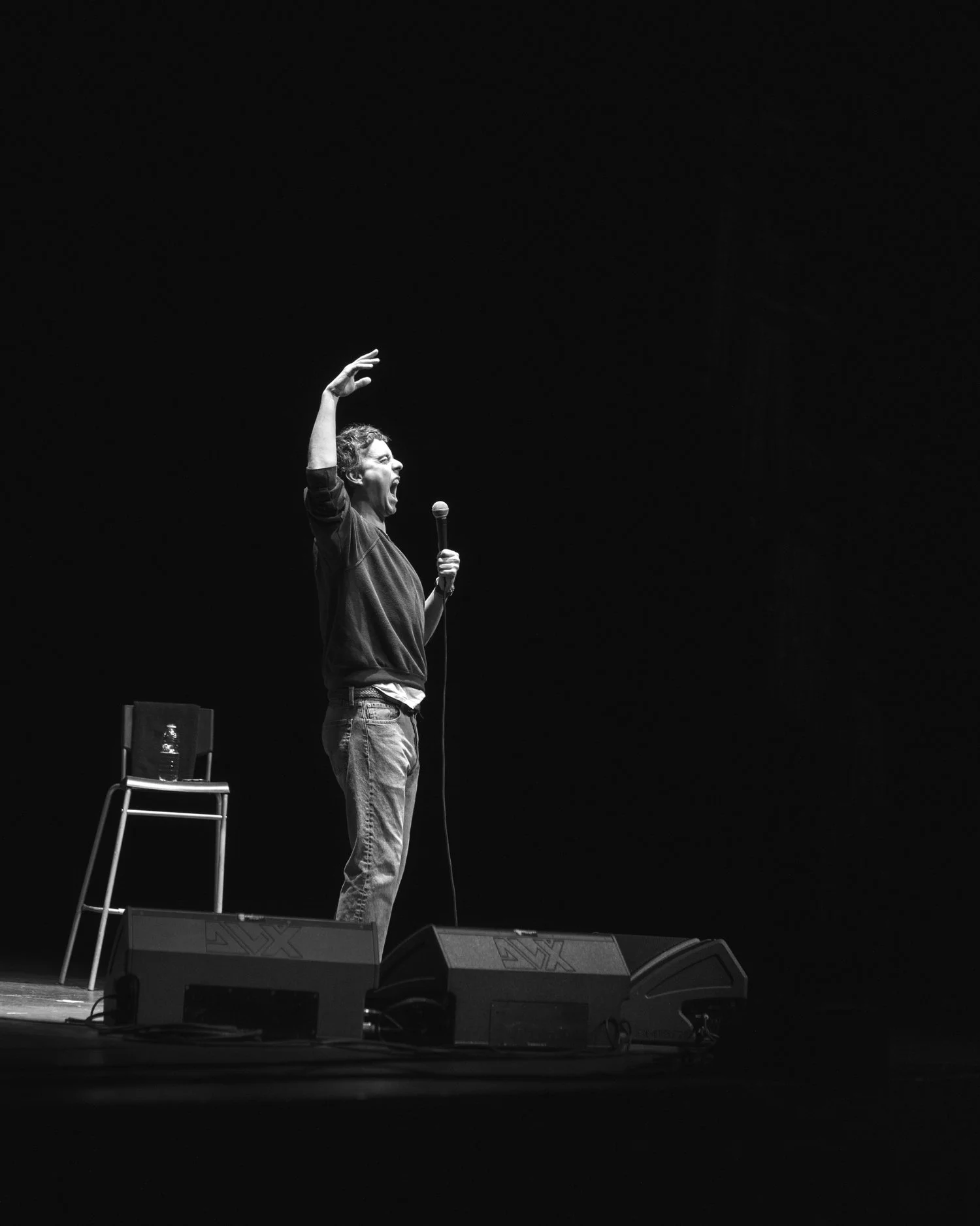 A person performs on stage, holding a microphone, with an animated expression and raised hand, in front of a dark background.