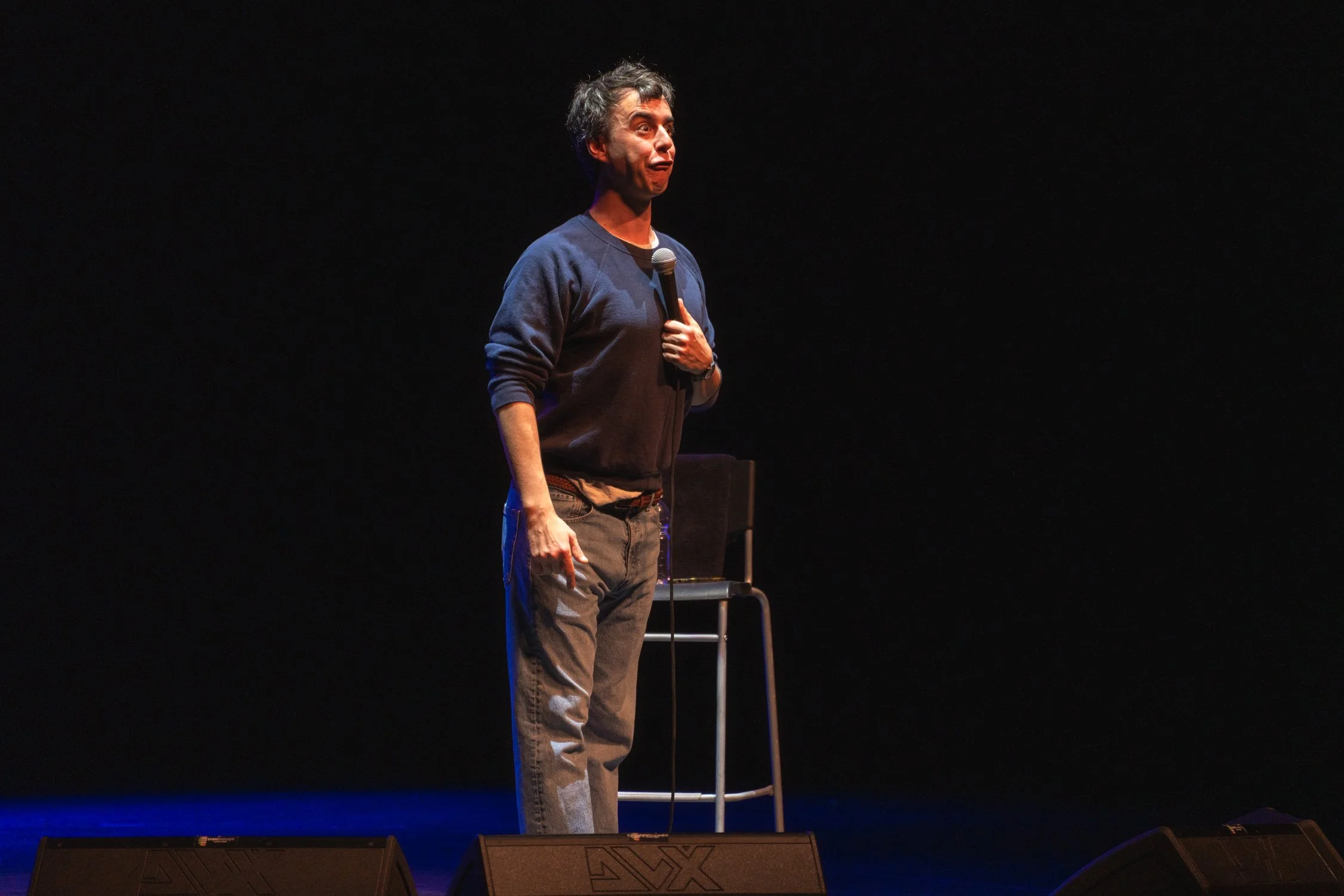 A man stands on a stage in front of a black background, holding a microphone and making a humorous facial expression.