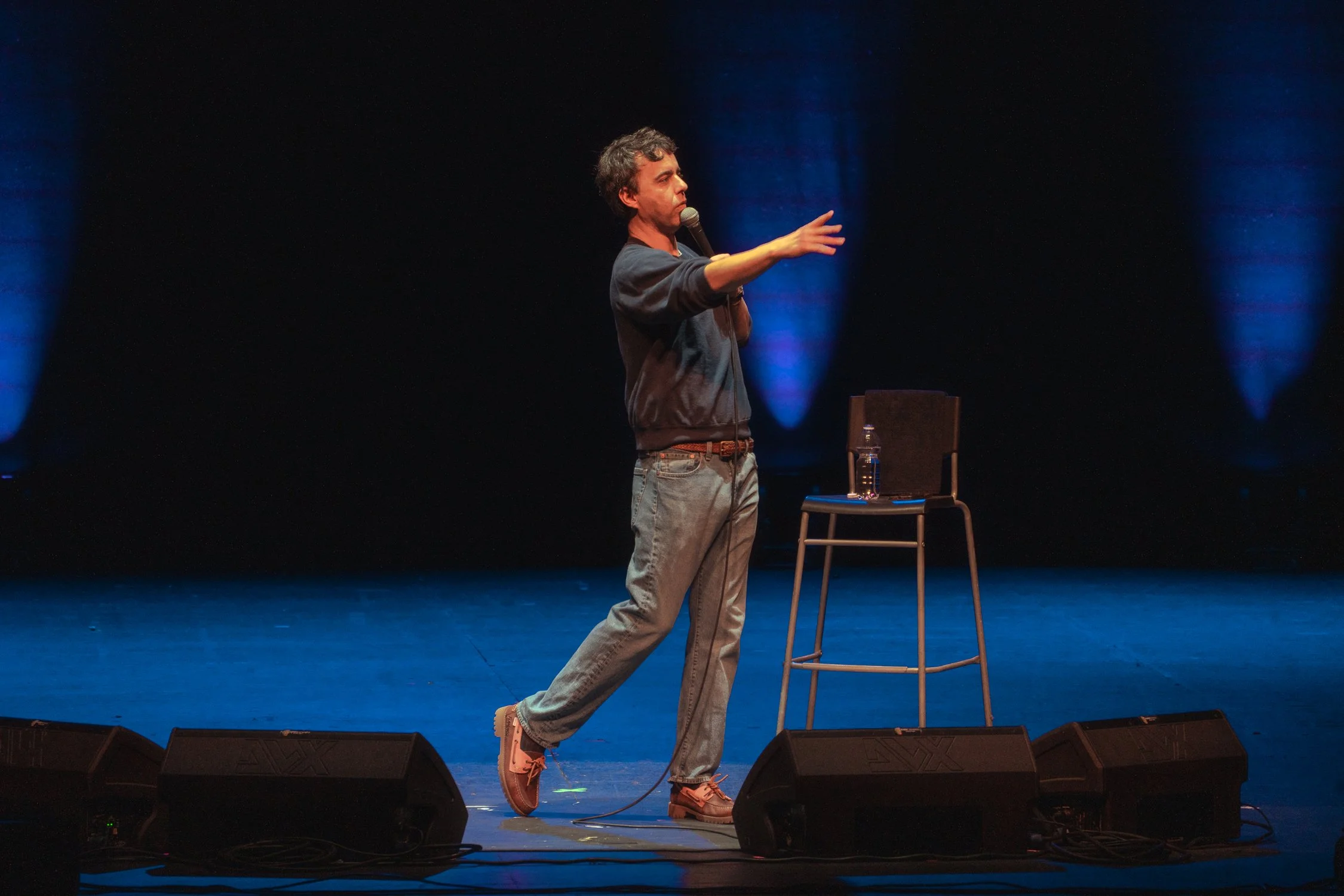 A man with curly dark hair and a beard stands on a stage, speaking into a microphone, gesturing with his right arm extended. He wears a dark long-sleeve shirt, light jeans, and brown shoes. A chair with a water bottle on it is next to him, and there 