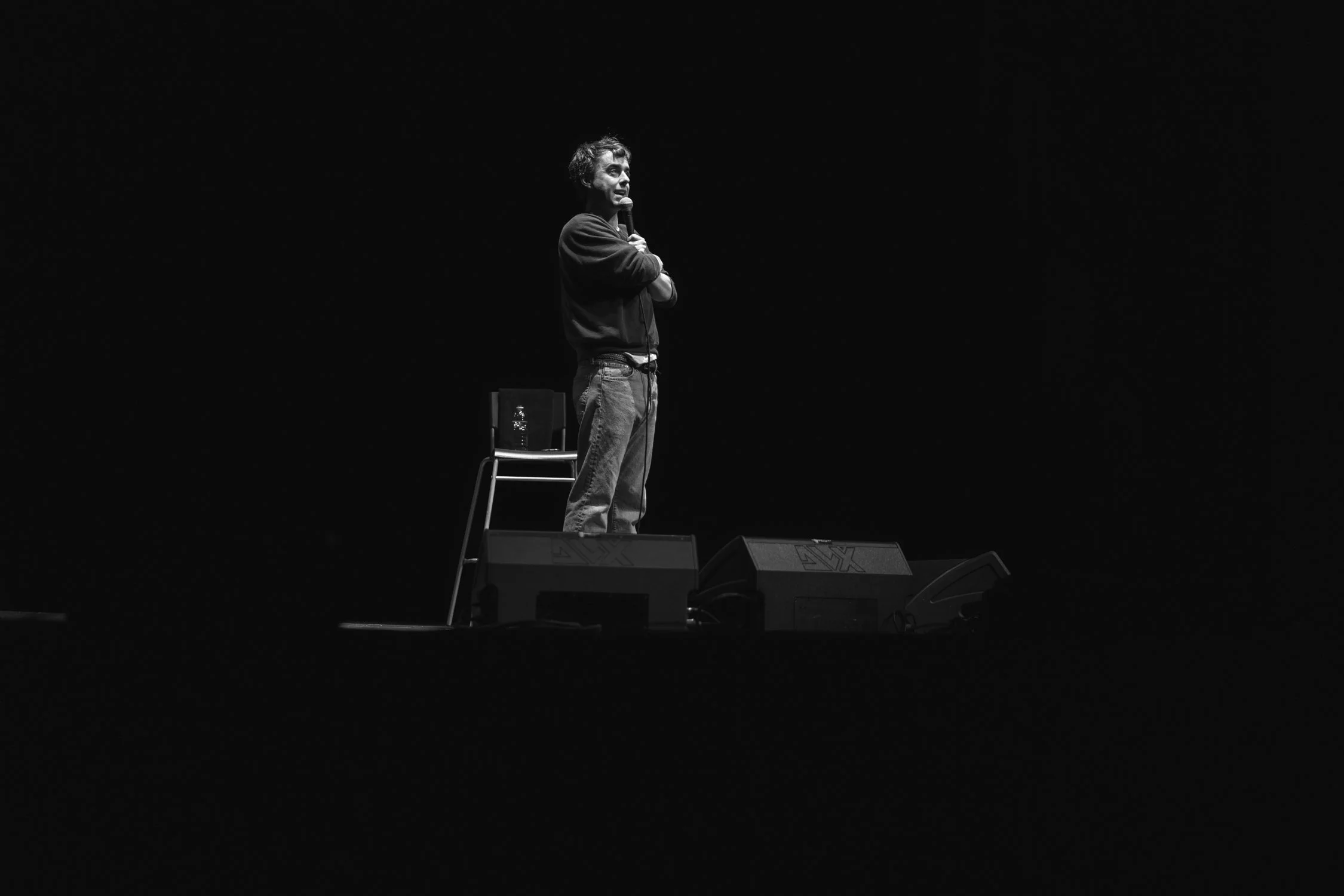 A man holding a microphone on stage, standing next to a chair with a bottle of water, in a dark environment with minimal lighting.