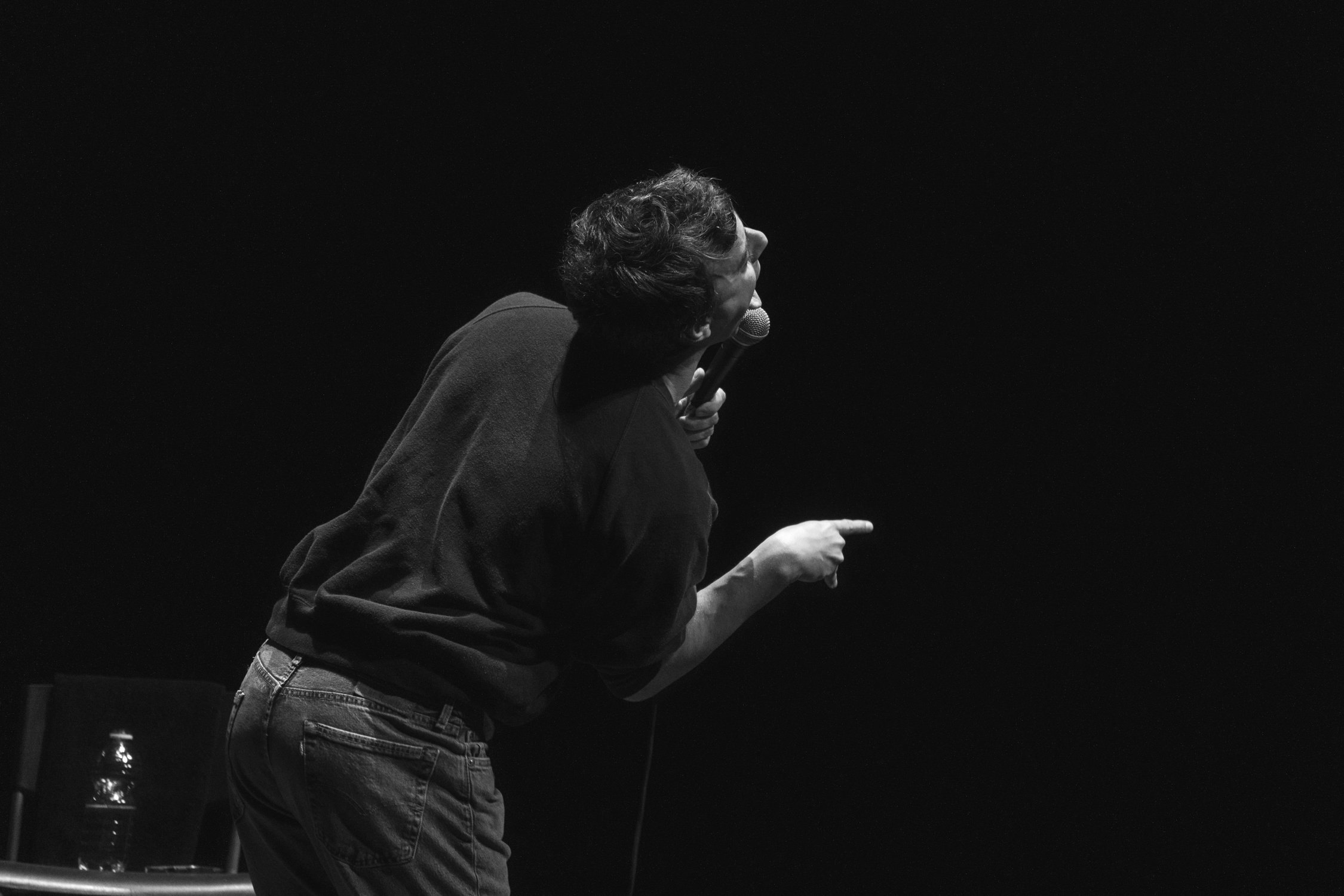 A person holding a microphone and pointing, seen from the side, against a dark background.