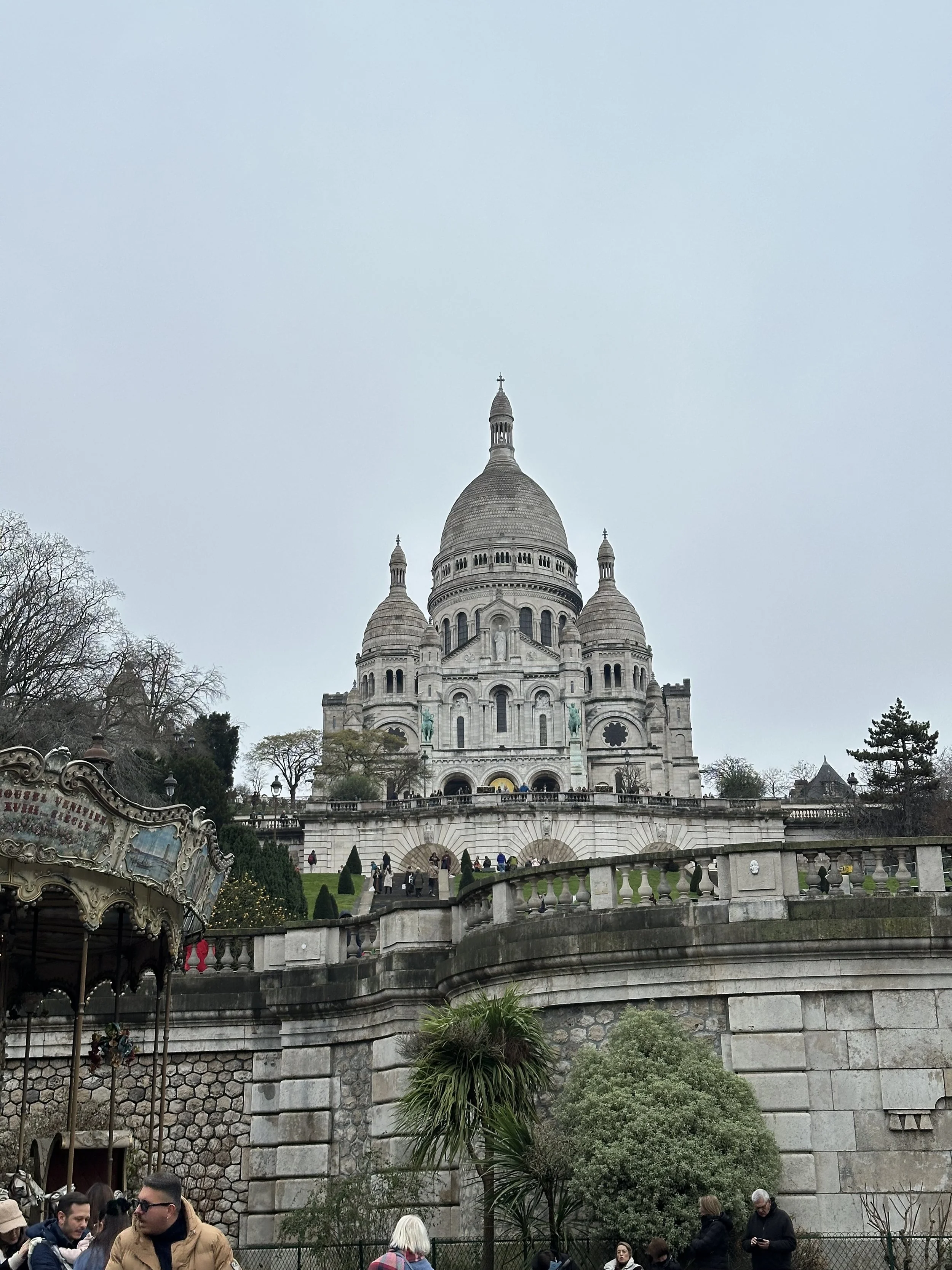 The Sacré-Cœur Basilica in Paris, France, seen from the front, with visitors and a carousel in the foreground, set on a cloudy day.