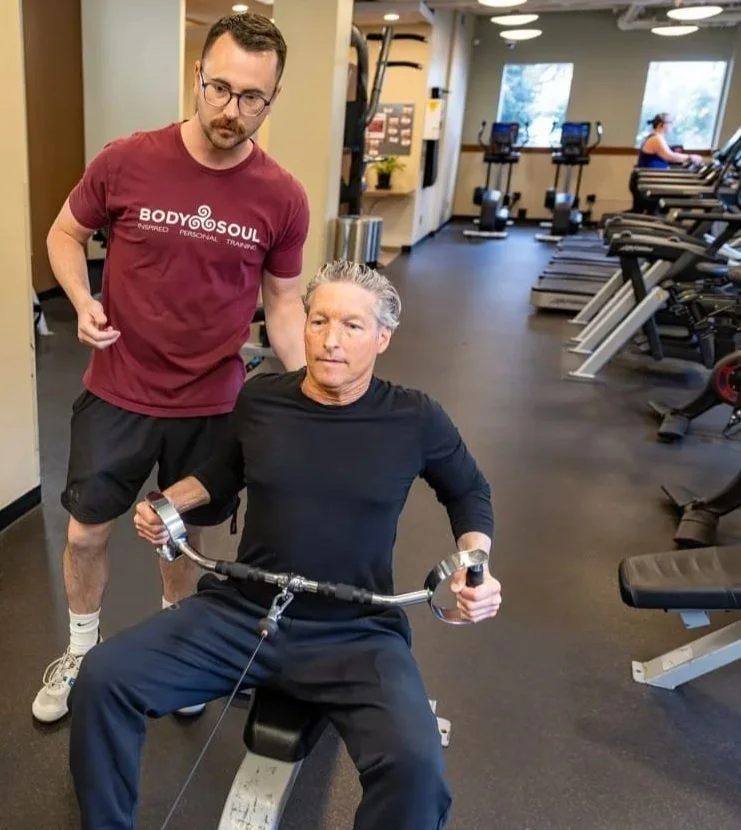 A man in a black shirt is sitting on a workout bench, holding a cable machine handle, preparing for exercise, while a trainer in a maroon shirt supervises him in a gym with treadmills and exercise equipment in the background.