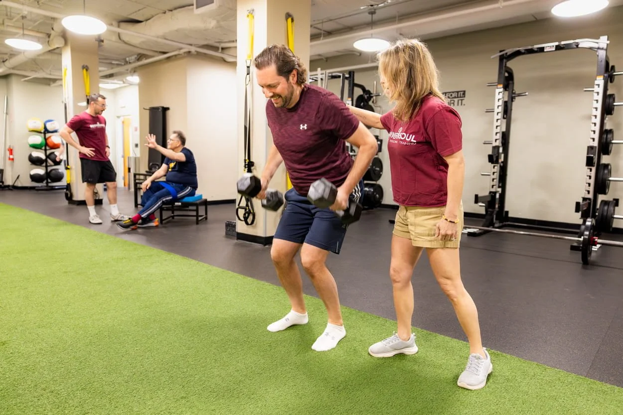 A man performs a fitness exercise with dumbbells assisted by a trainer in a gym. In the background, another person sits and interacts with a trainer. The gym has weight racks, exercise equipment, and a green turf area.