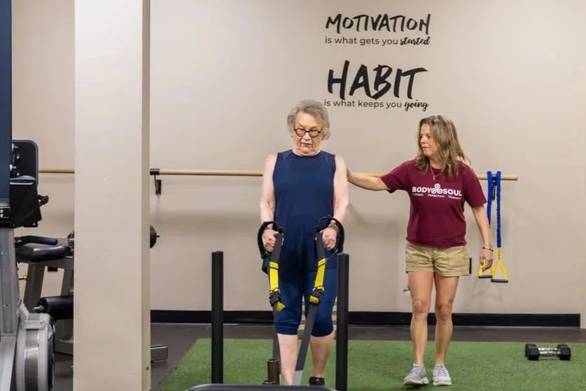 An elderly woman in workout clothes using resistance bands attached to a harness, assisted by a trainer in a gym with motivational quotes on the wall.