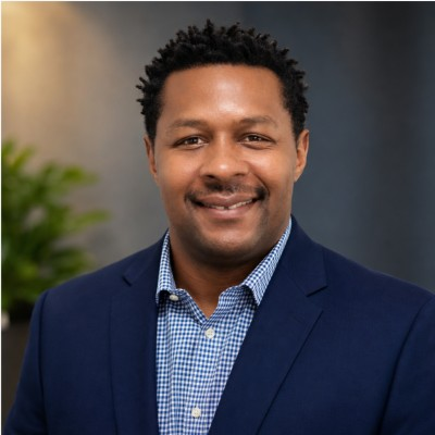A black man with short slighly spikey hair. He is smiling and looking at the camera and is wearing a blue blazer and light blue and white checked button up shirt. 