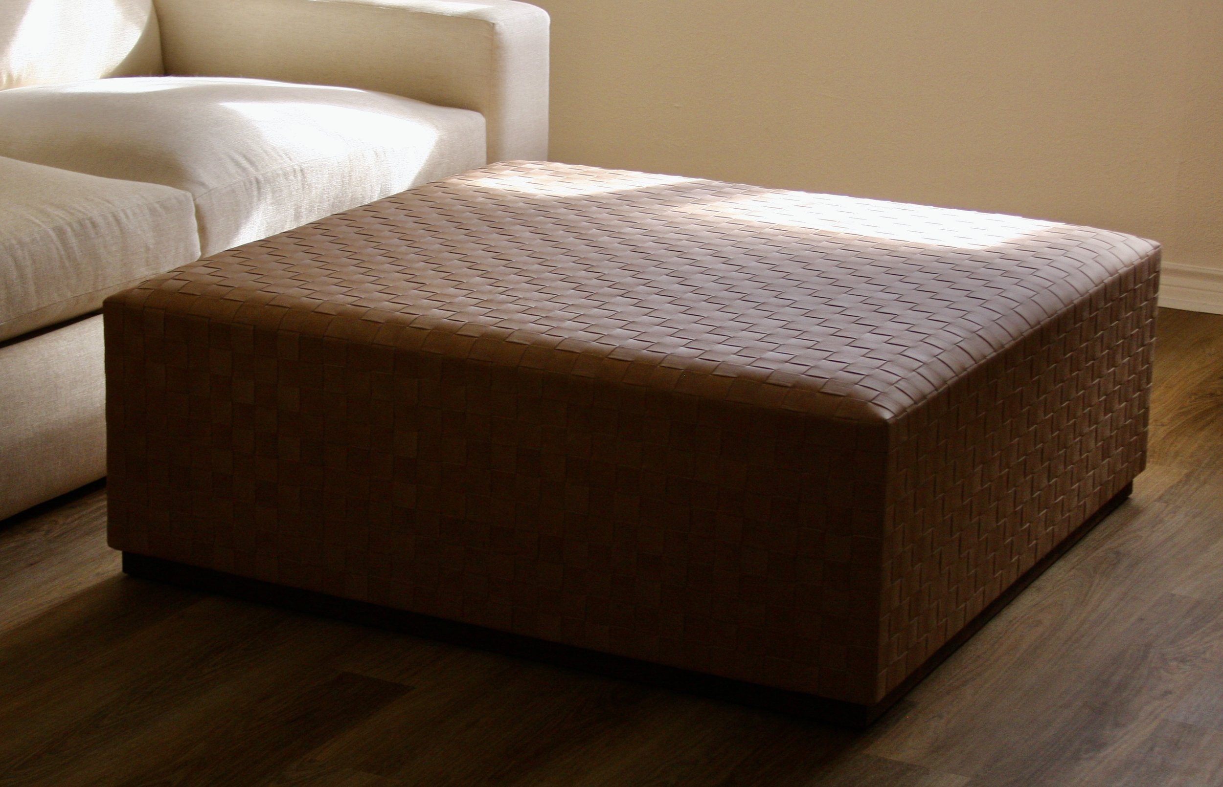 A woven ottoman in front of a beige sofa on wooden flooring, with sunlight casting shadows on the furniture.