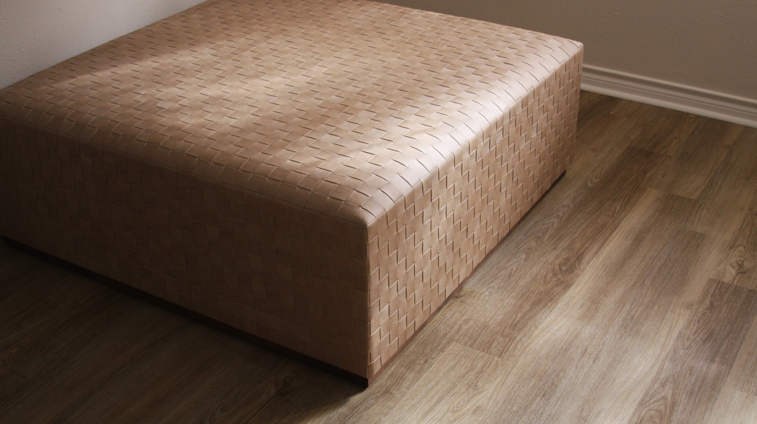 A square, upholstered ottoman with a woven pattern in a light brown or tan color, placed on a wooden floor next to a wall with trim.