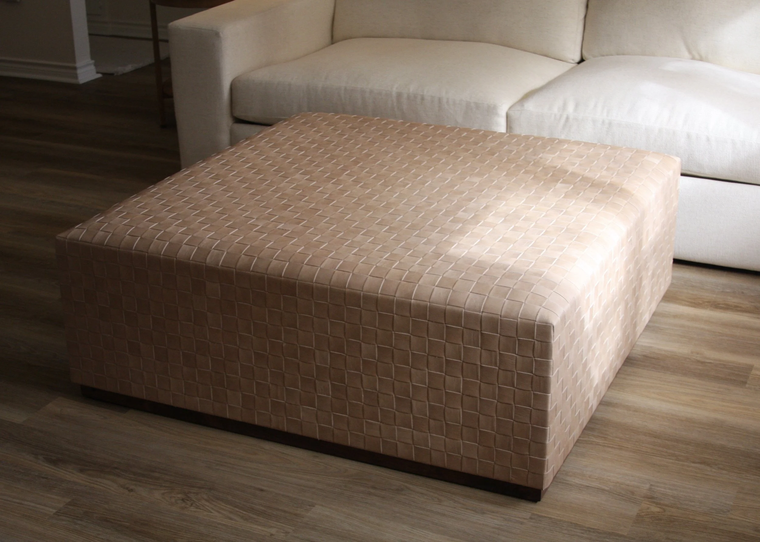 A beige, textured square ottoman in front of a white sofa on wooden flooring.