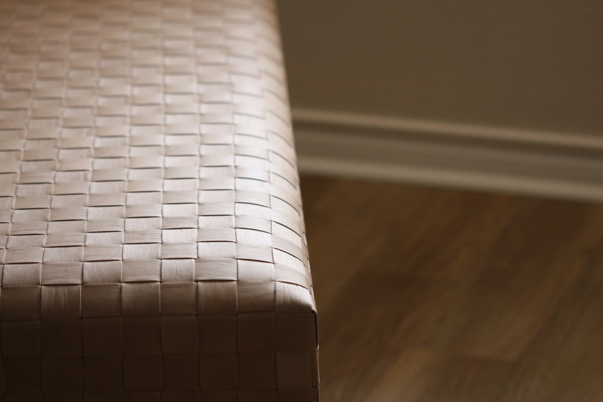 Close-up of a woven wooden or wicker bench with a blurred wooden floor and wall in the background.
