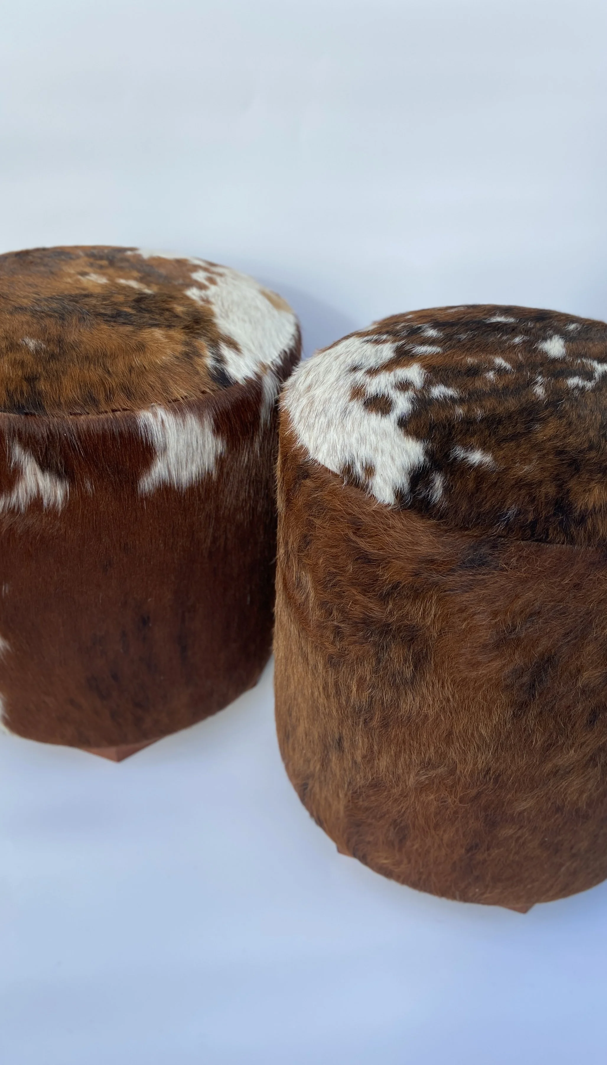 Two cylindrical brown and white cowhide stools with fur texture on a white background.