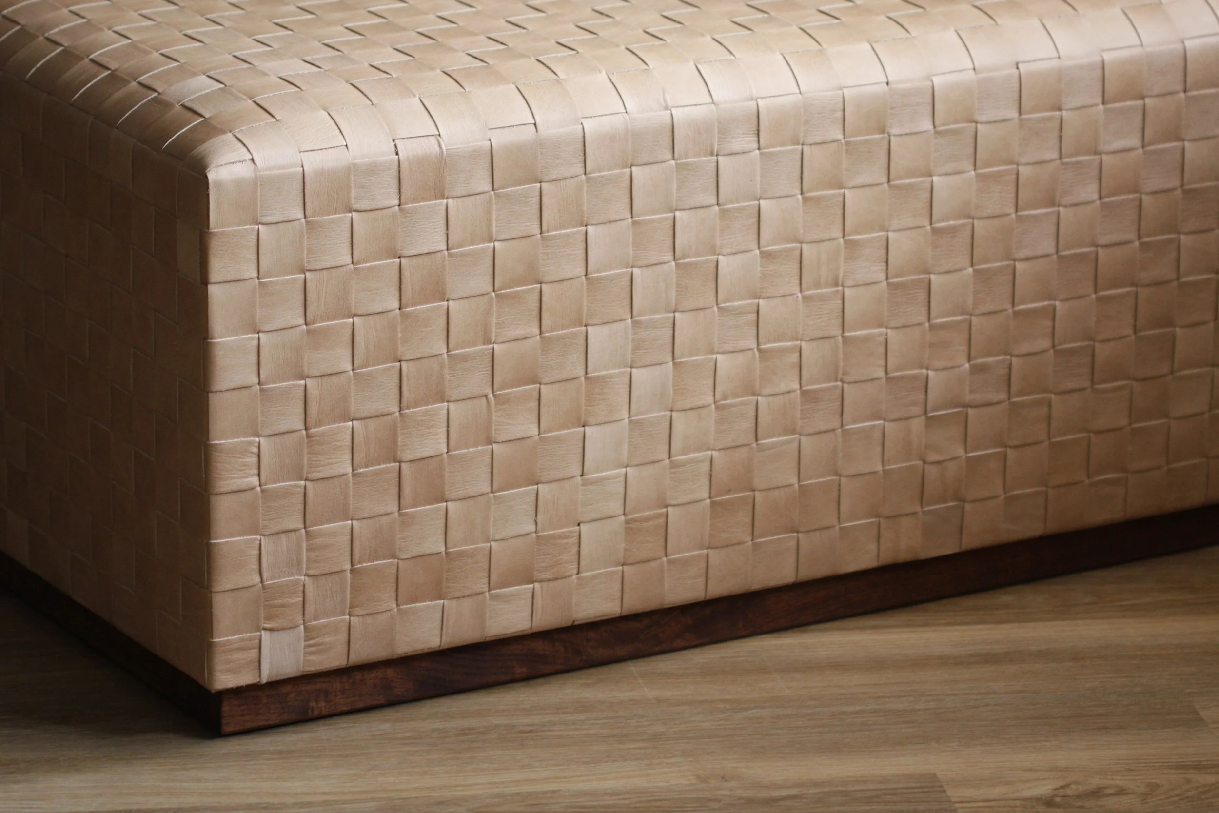 Close-up of a woven beige leather ottoman with a wooden base on a wooden floor.