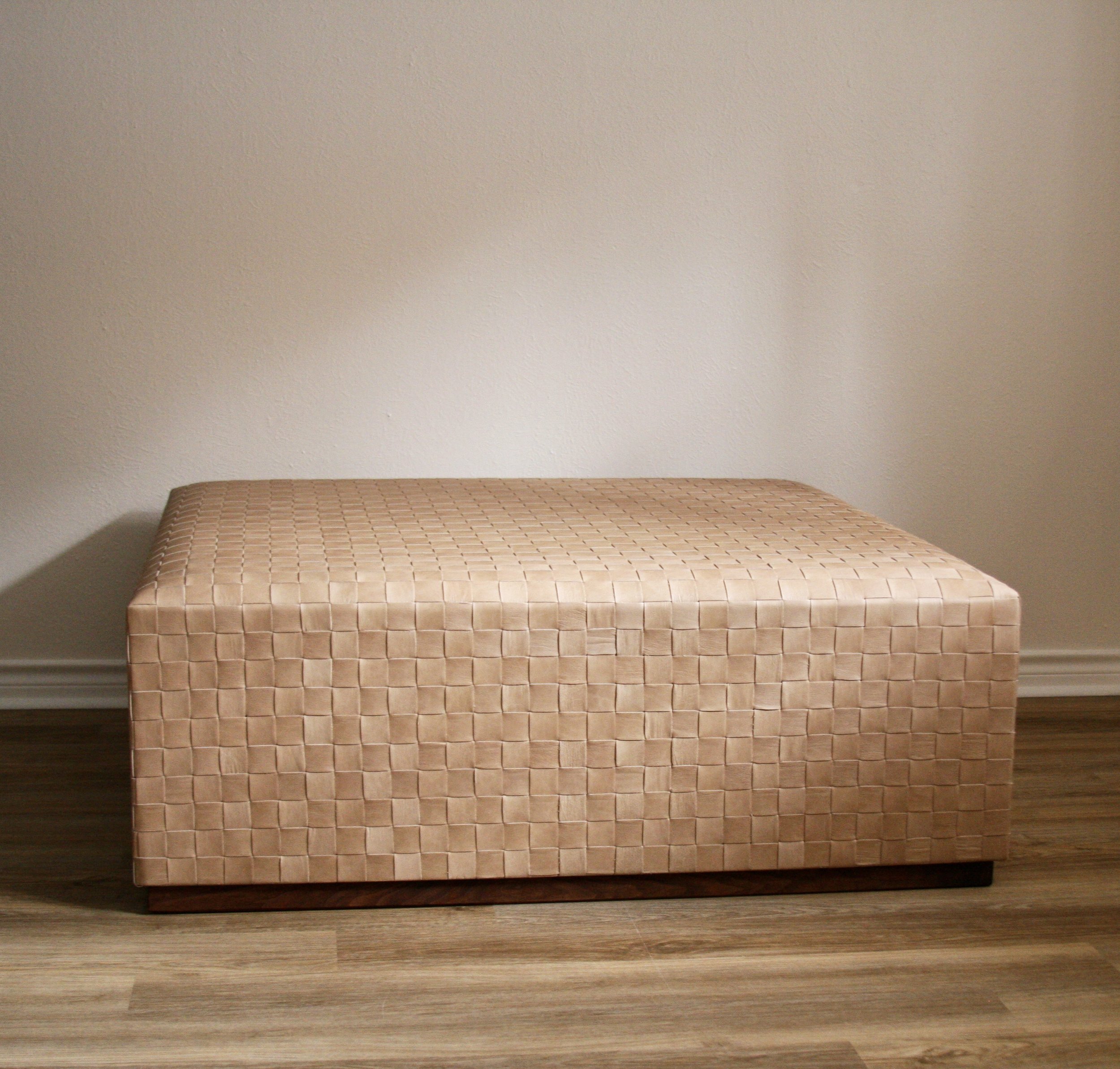A rectangular beige woven-pattern upholstered storage bench against a plain white wall, on a wooden floor.