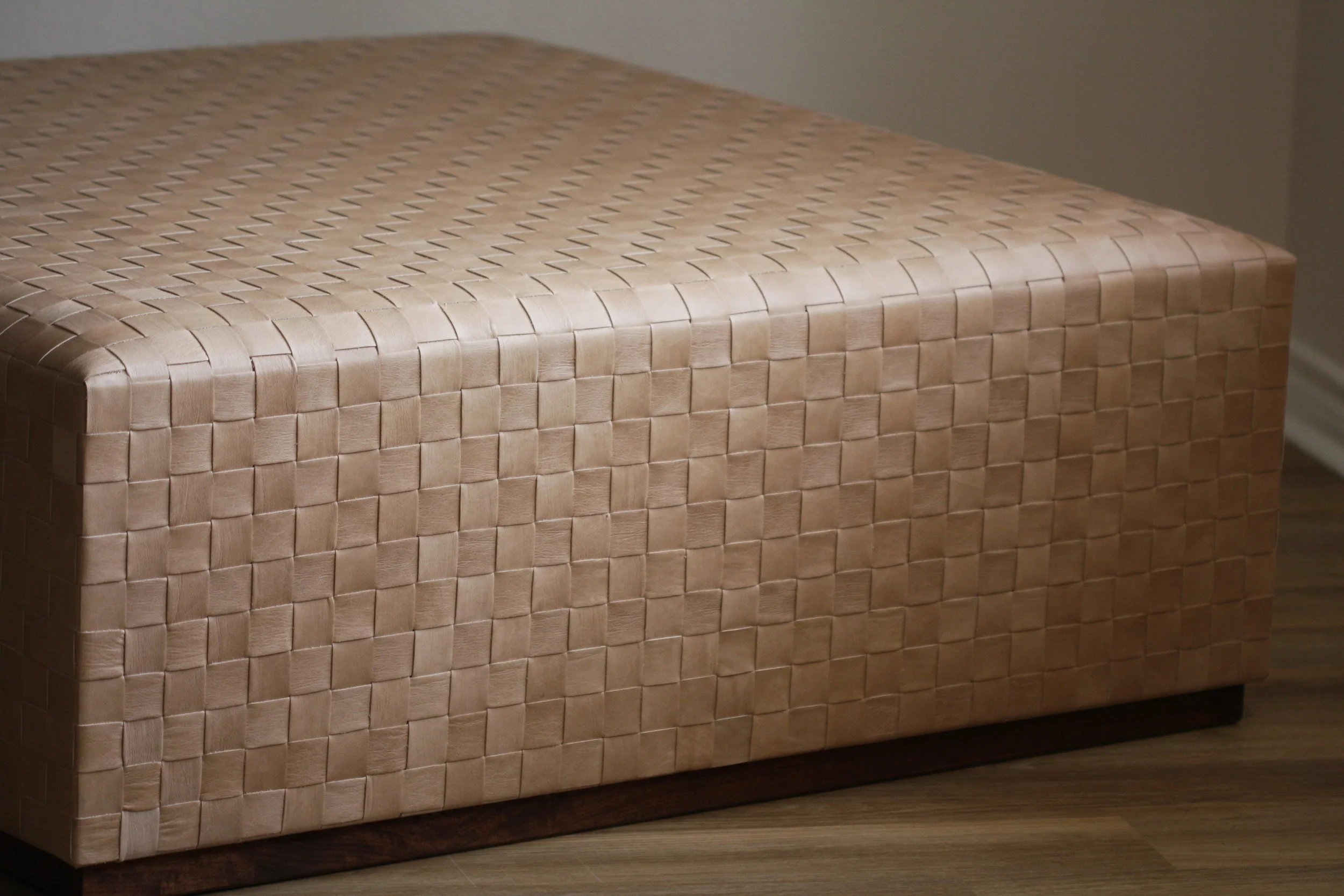 A rectangular woven leather ottoman with a light tan color, placed on a wooden floor against a beige wall.
